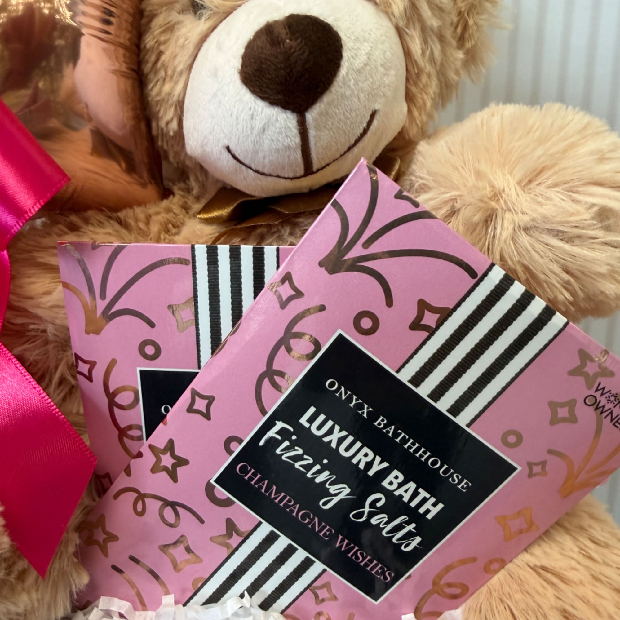 Two pink bath salt packages beside a teddy bear with a ribbon.