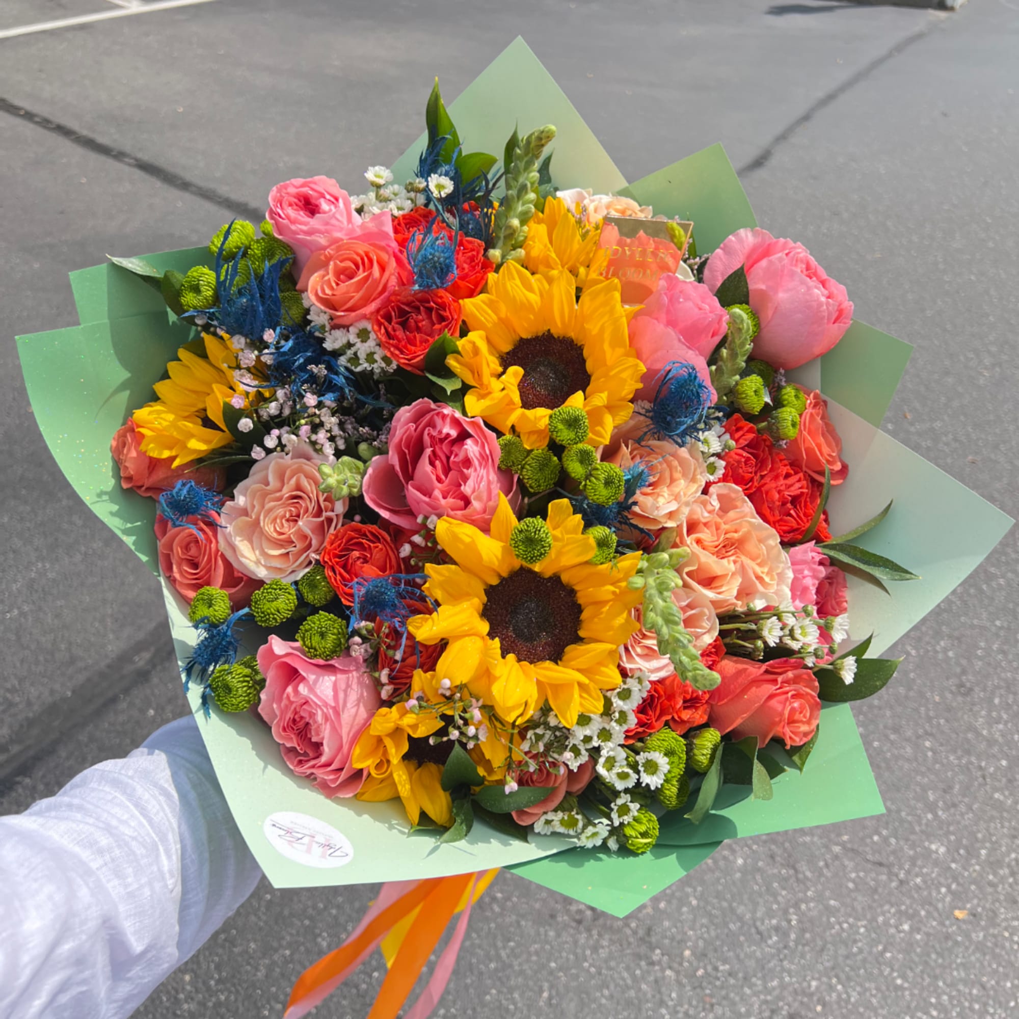 Handheld bouquet of sunflowers, roses, and mixed blooms wrapped in green paper