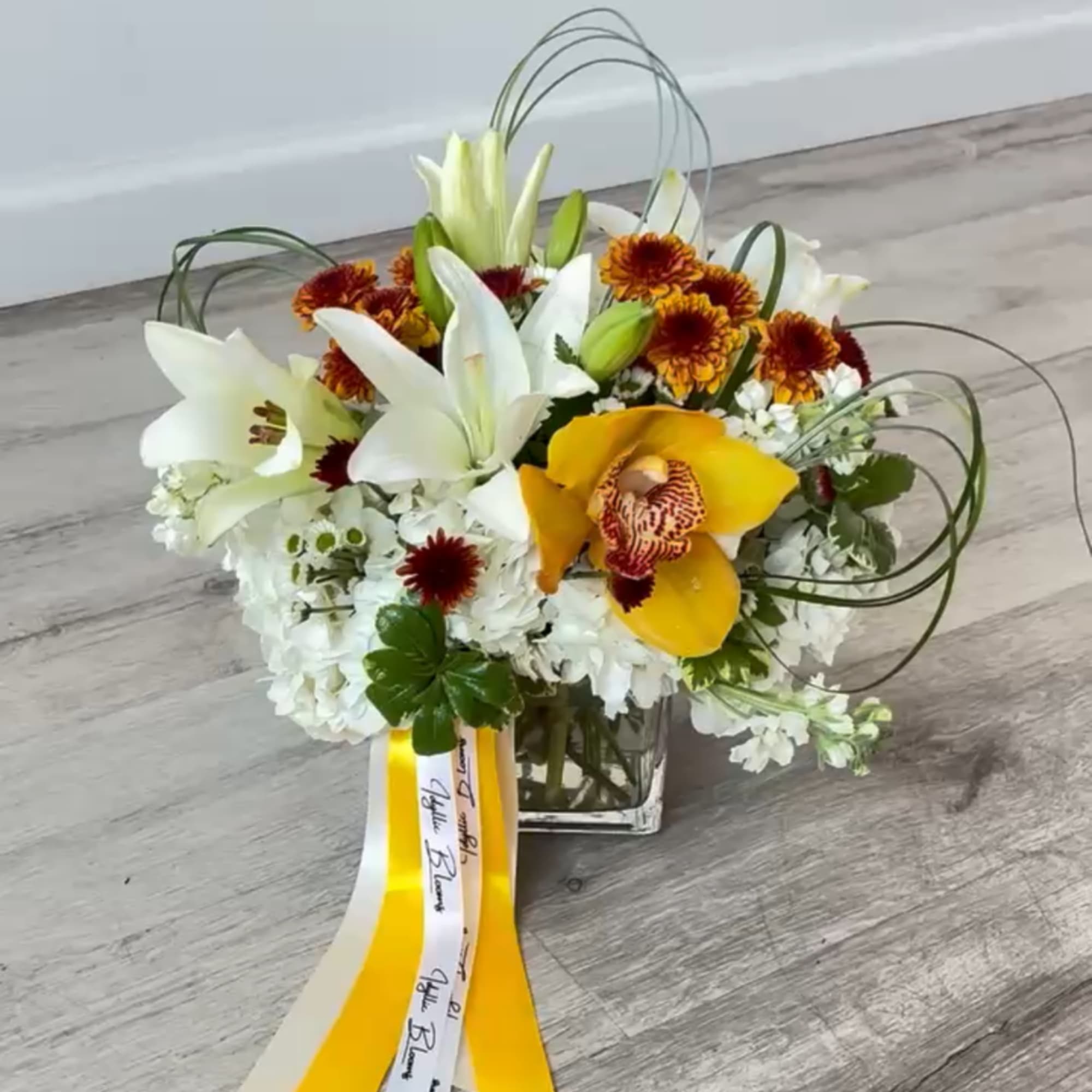 White lilies and yellow orchids in a glass vase with ribbon tails