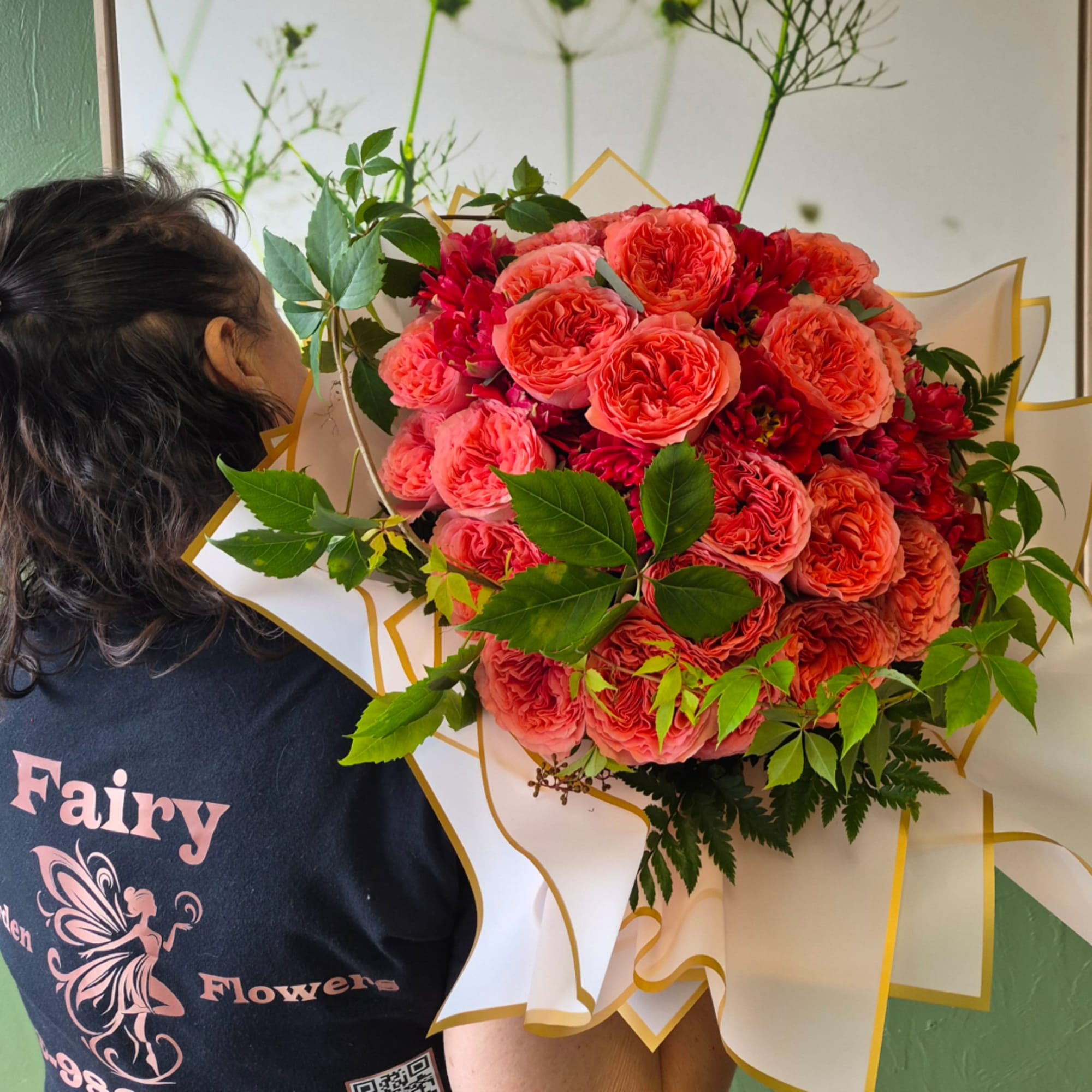 Person holding a bouquet of coral and pink roses wrapped in white paper with gold trim