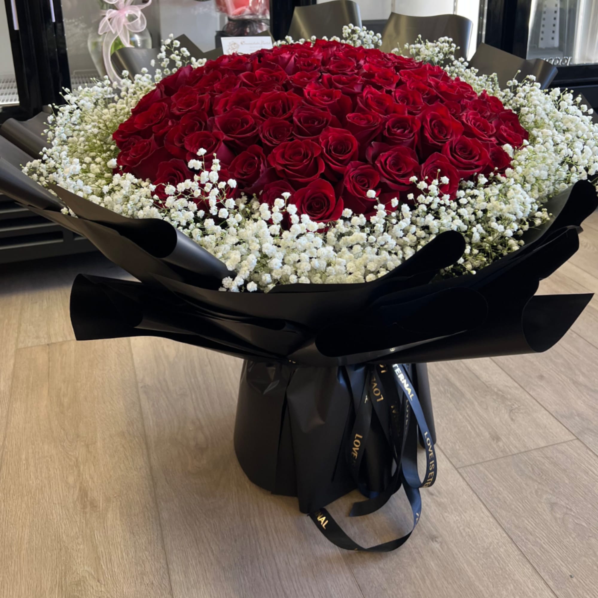 Large round bouquet of red roses encircled by white filler flowers in glossy black wrapping.