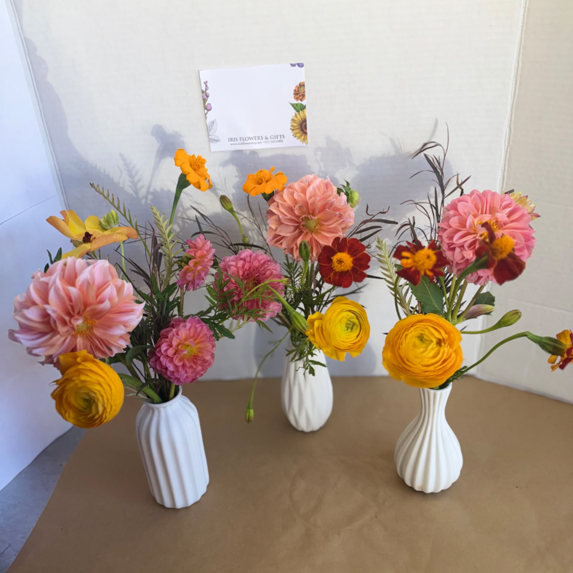 Three small floral arrangements in white vases with pink, yellow, and orange blooms