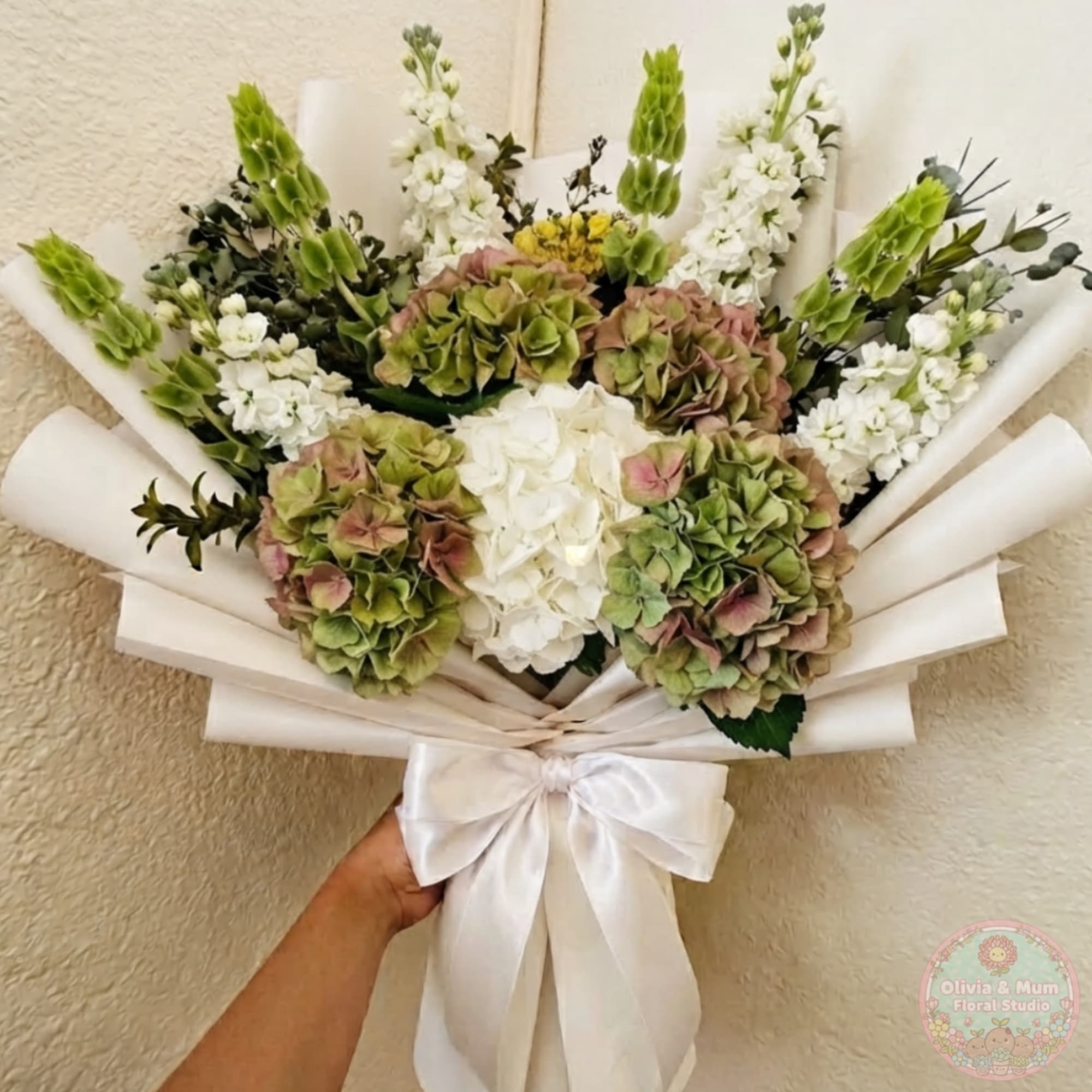 Handheld bouquet of white and green flowers wrapped in white paper with a ribbon