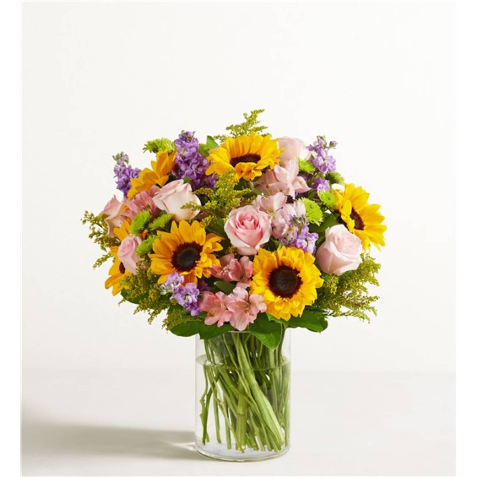 Mixed bouquet of sunflowers, pink roses, and purple flowers in a clear glass vase
