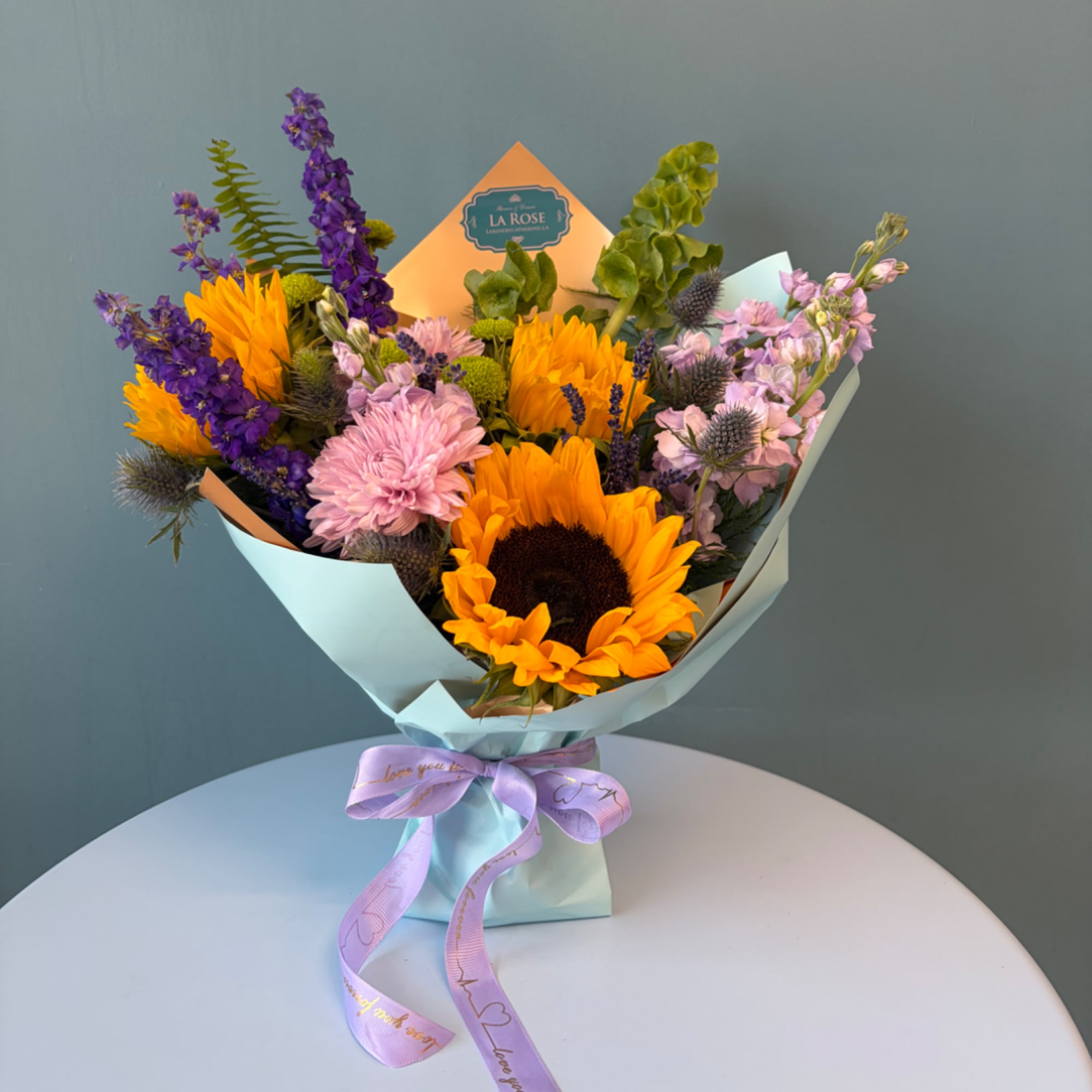 Bouquet of sunflowers, purple blooms, and pink flowers wrapped in pastel paper
