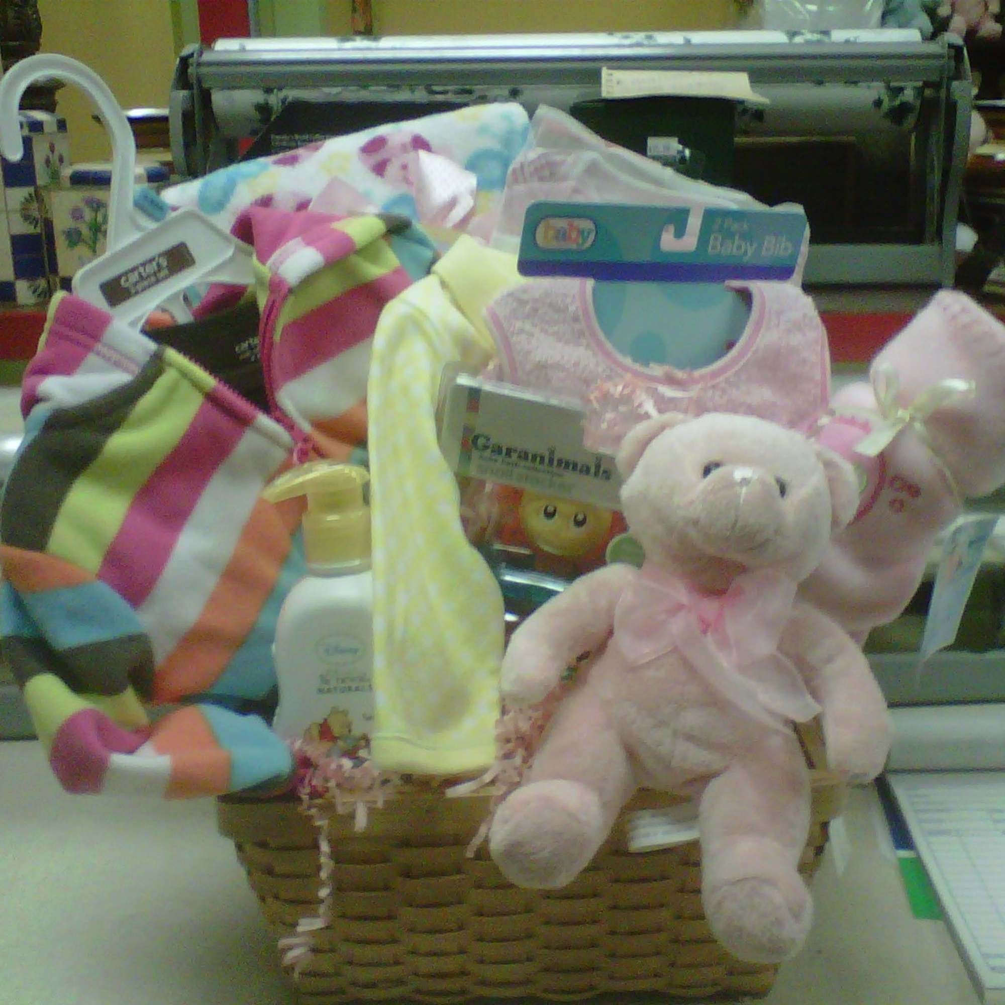 Baby gift basket with striped clothing, pink teddy bear, bibs, blanket, and baby lotion in a woven basket