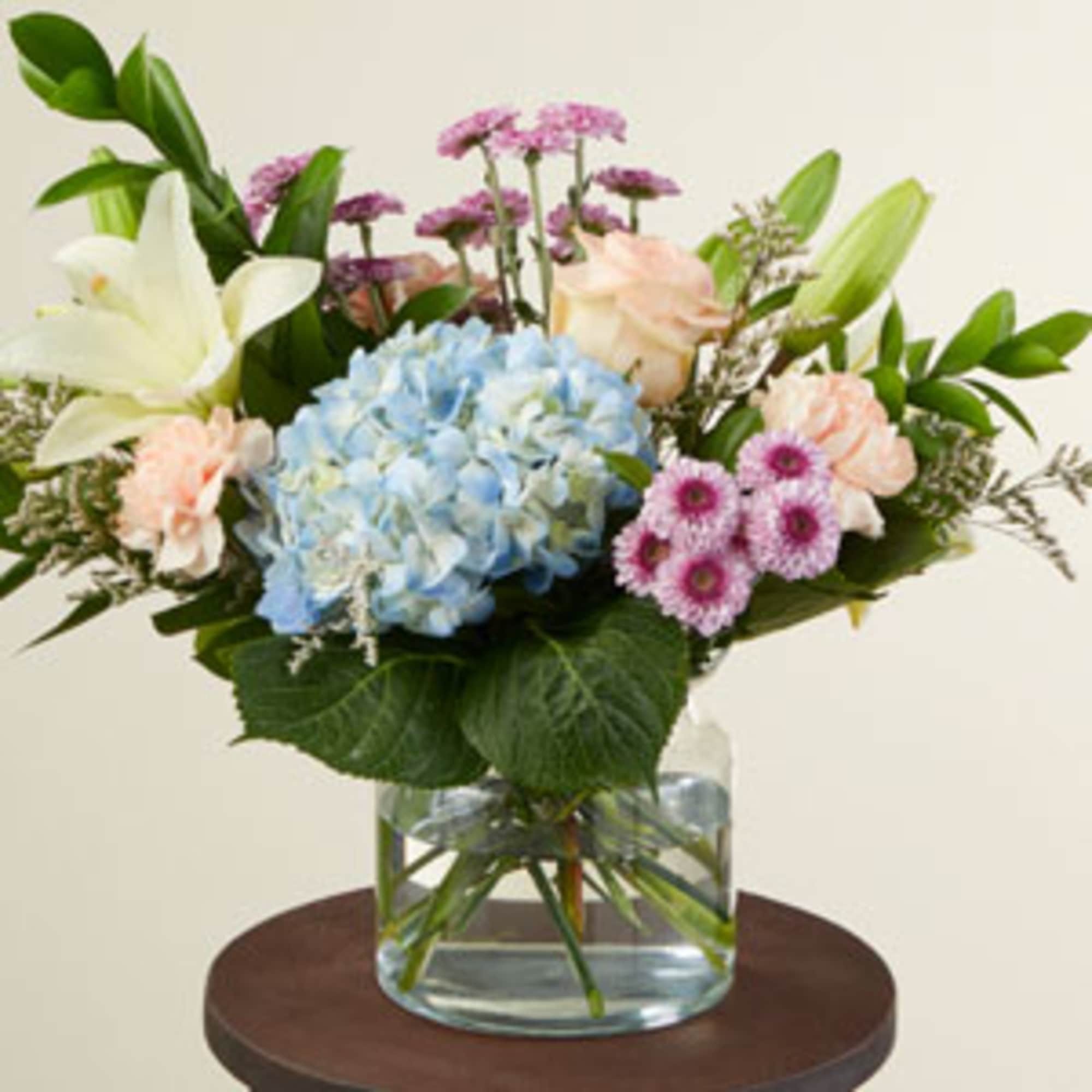 Mixed bouquet with blue hydrangea, lilies, roses, and pink daisies in a glass vase