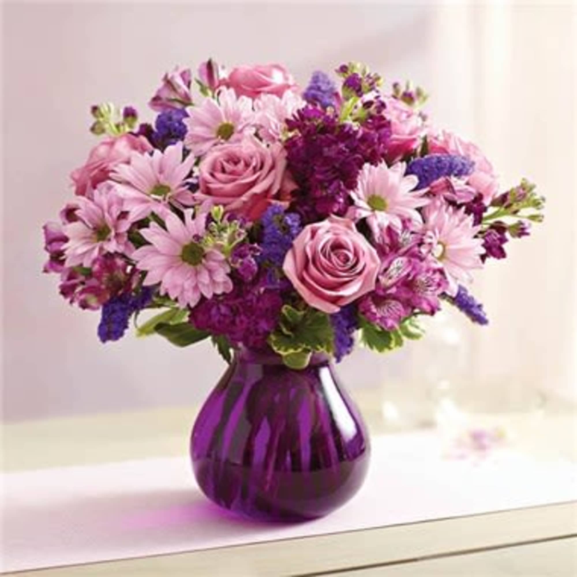 Pink and purple bouquet with roses and daisies arranged in a rounded purple glass vase