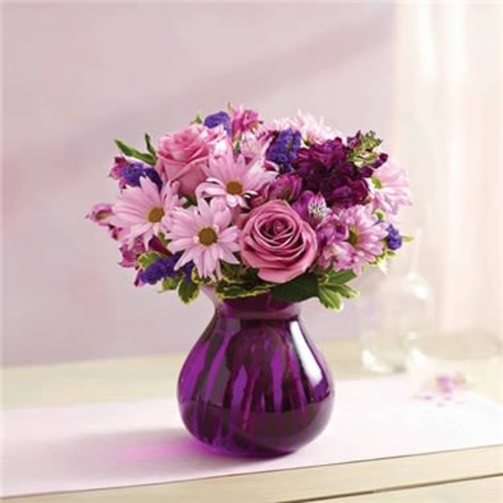 Pink roses and daisies with purple flowers arranged in a round purple glass vase