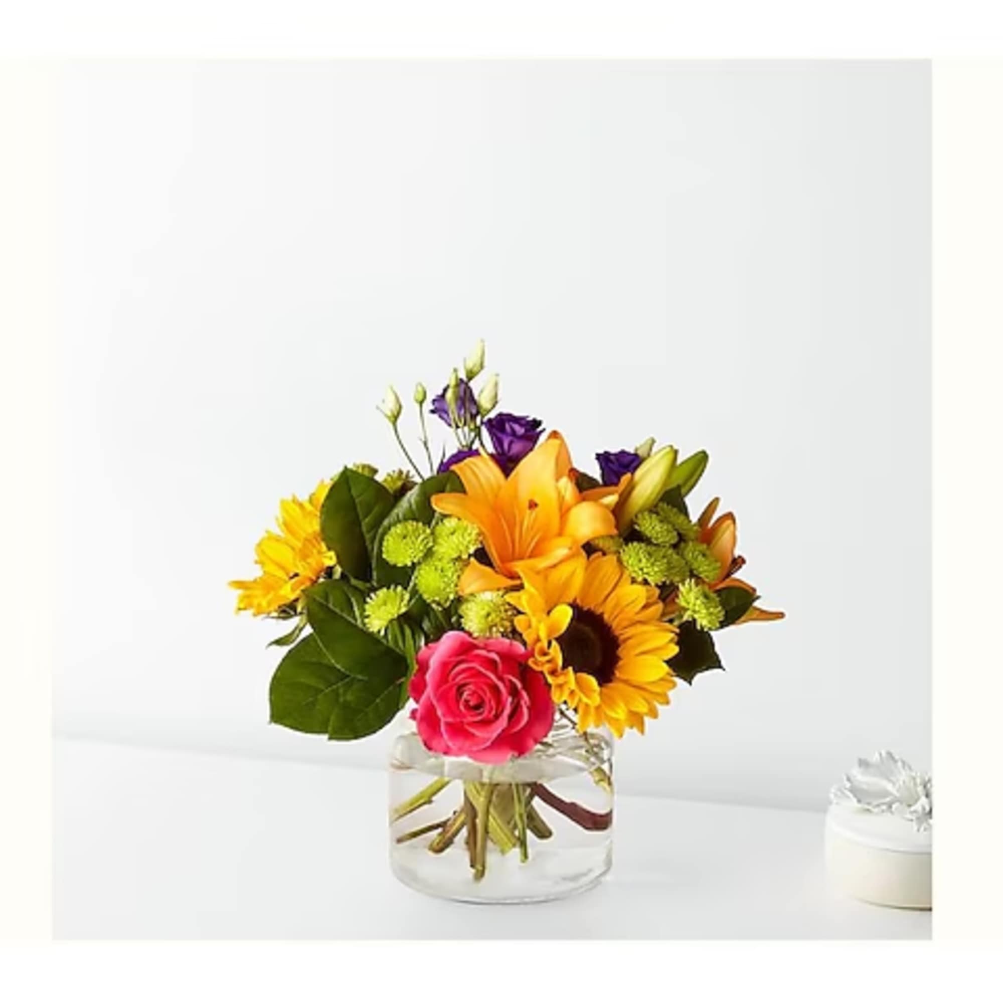 Bouquet of orange lilies, sunflowers, and a pink rose in a clear glass vase.