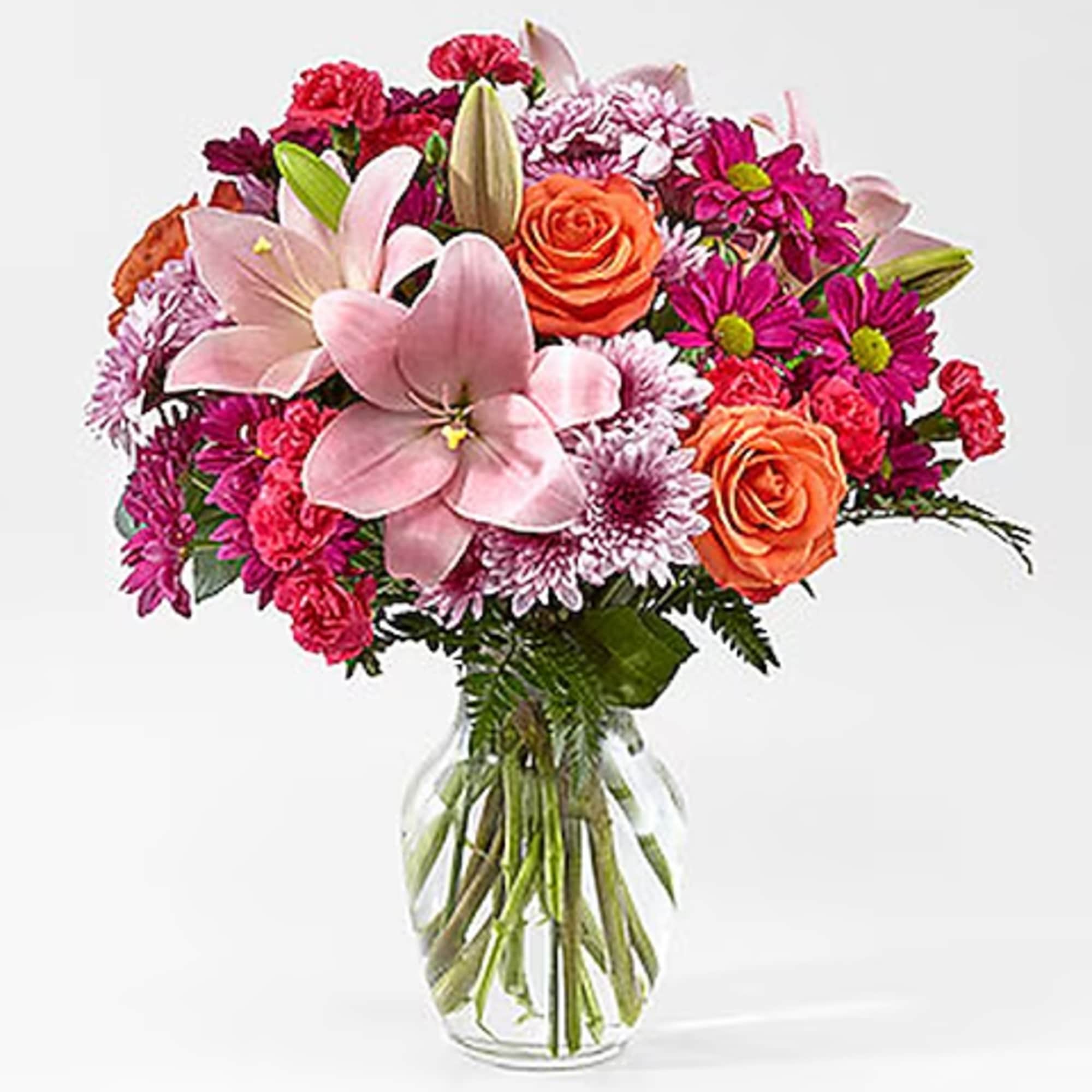 Mixed bouquet of pink lilies, orange roses, and magenta daisies in a clear glass vase