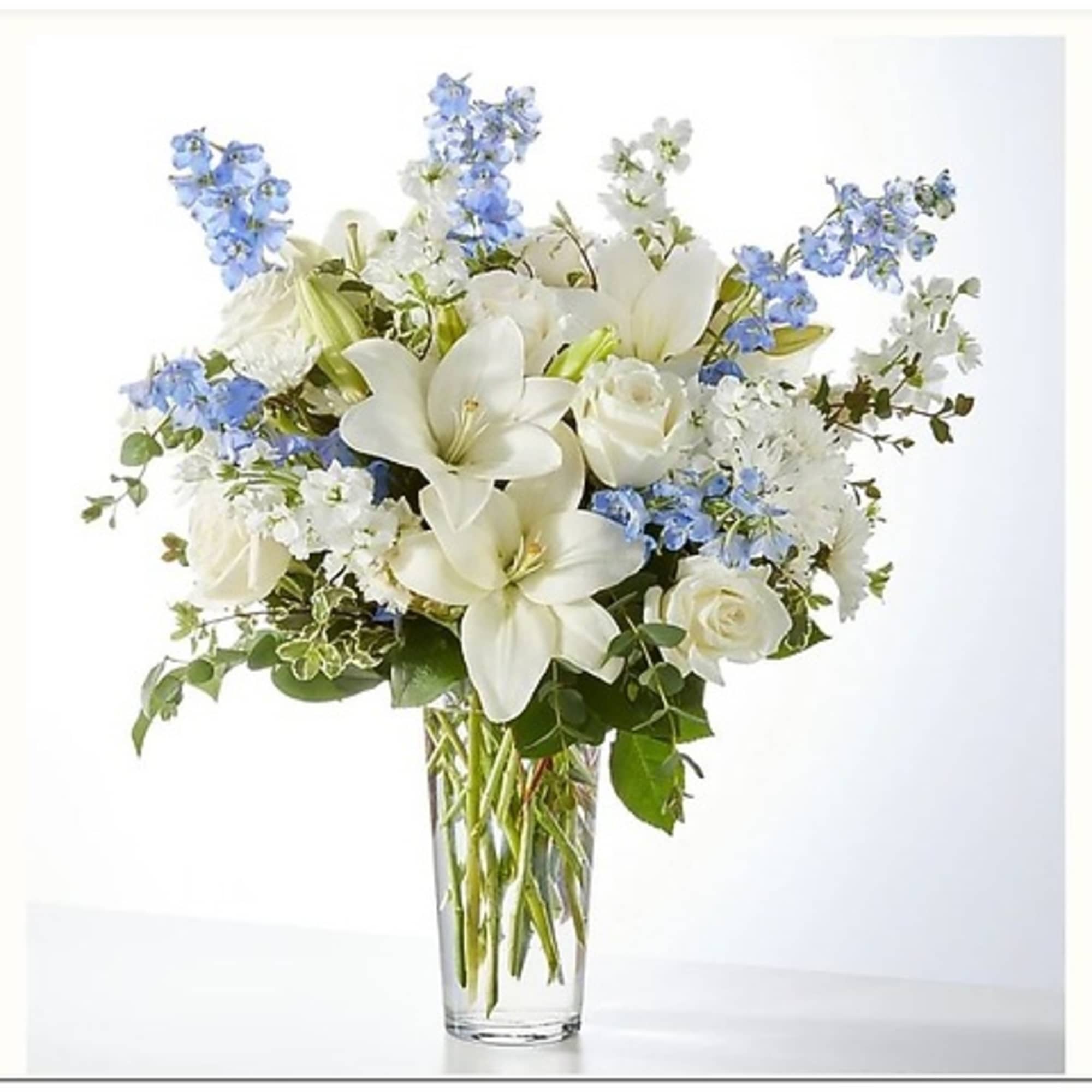 White lilies and roses with blue flowers in a clear glass vase