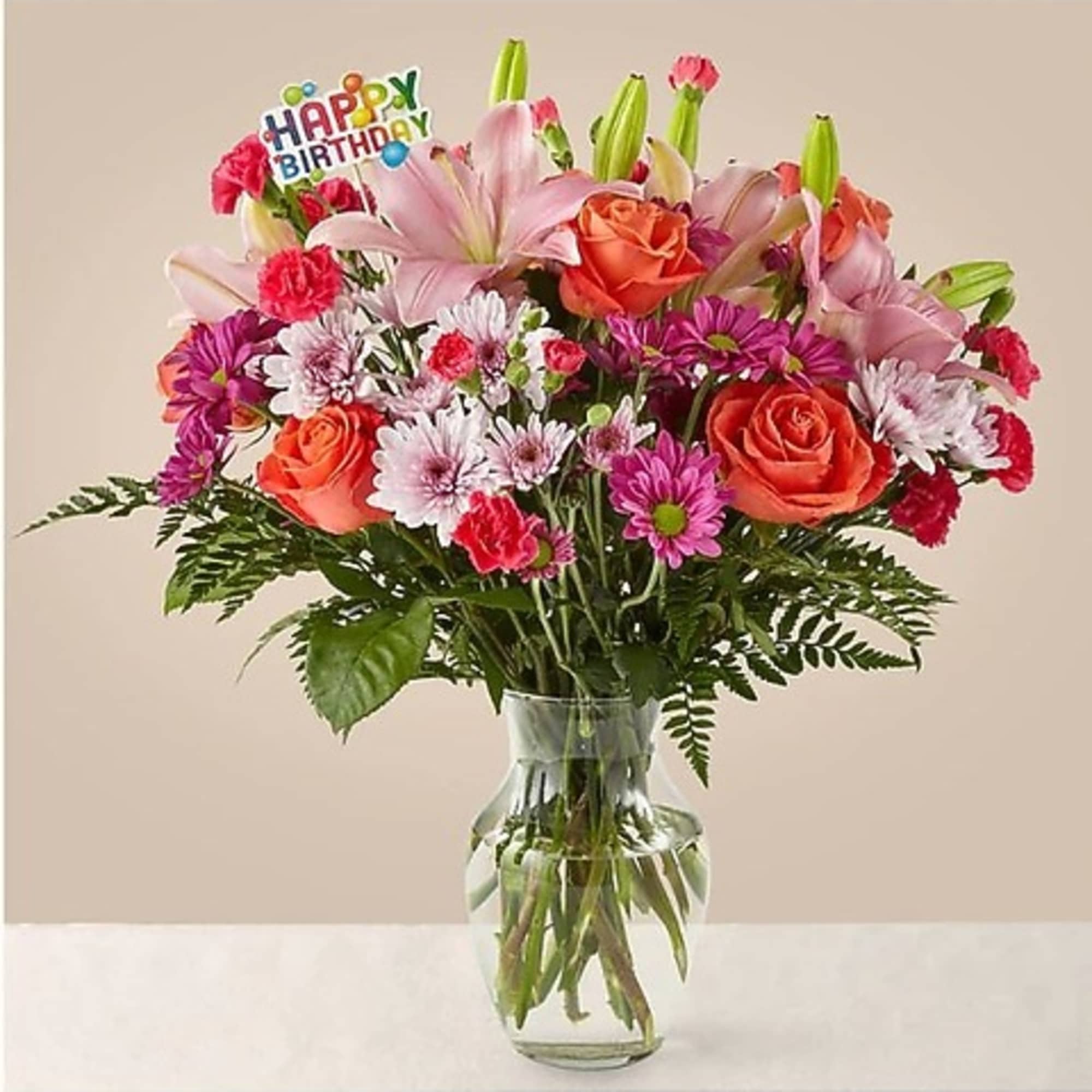 Mixed bouquet of pink lilies, roses, and daisies in a glass vase with a birthday pick