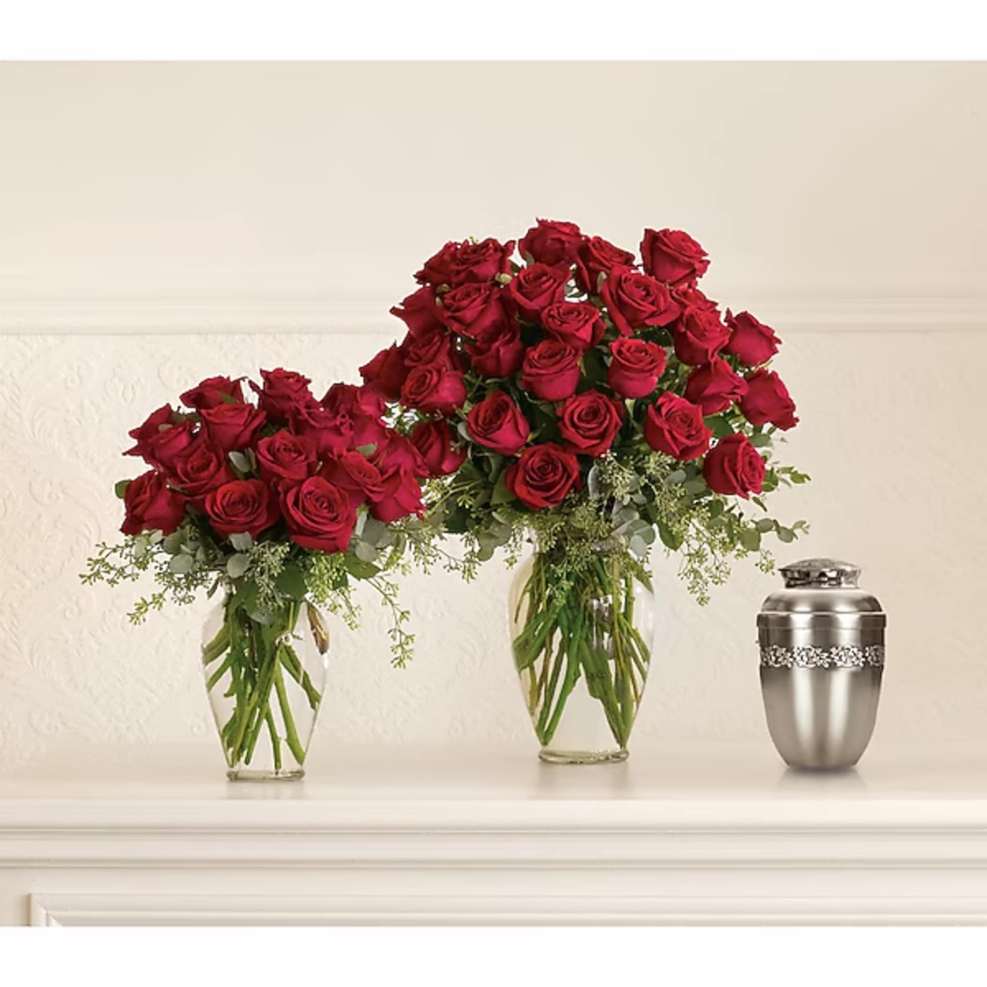 Two red rose arrangements in clear glass vases beside a silver urn