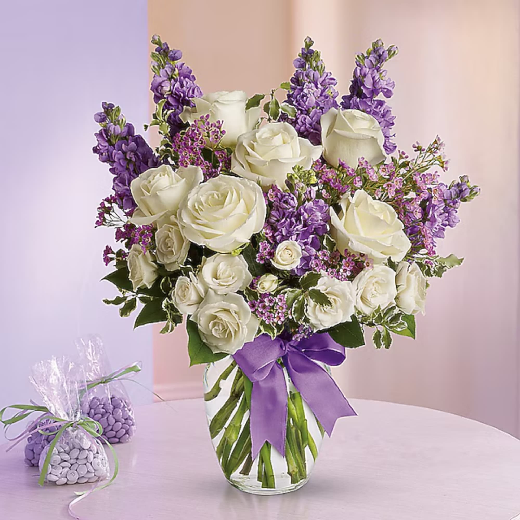 White roses and purple flowers arranged in a clear vase with a purple ribbon