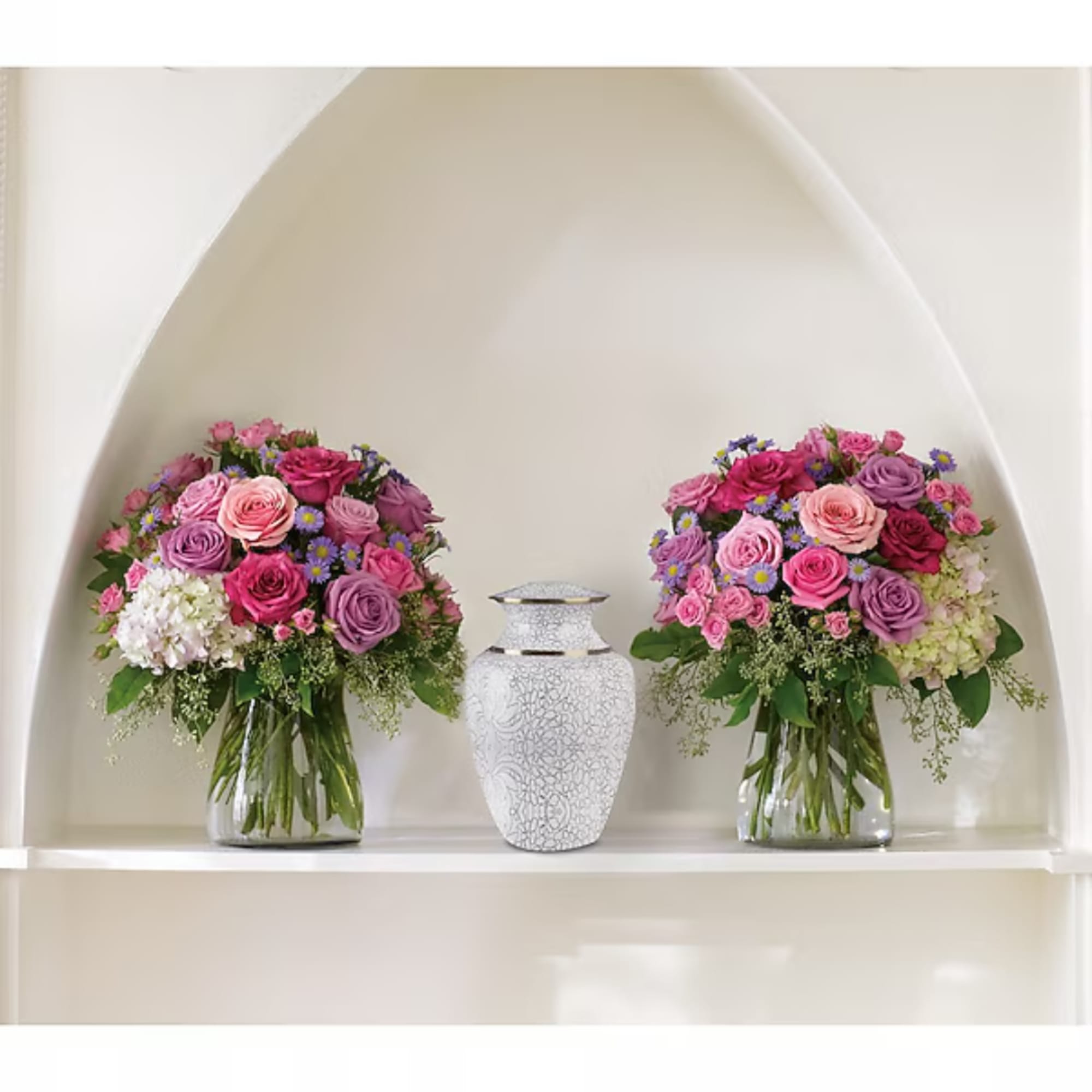Two pink and purple floral arrangements in glass vases with a white urn between them