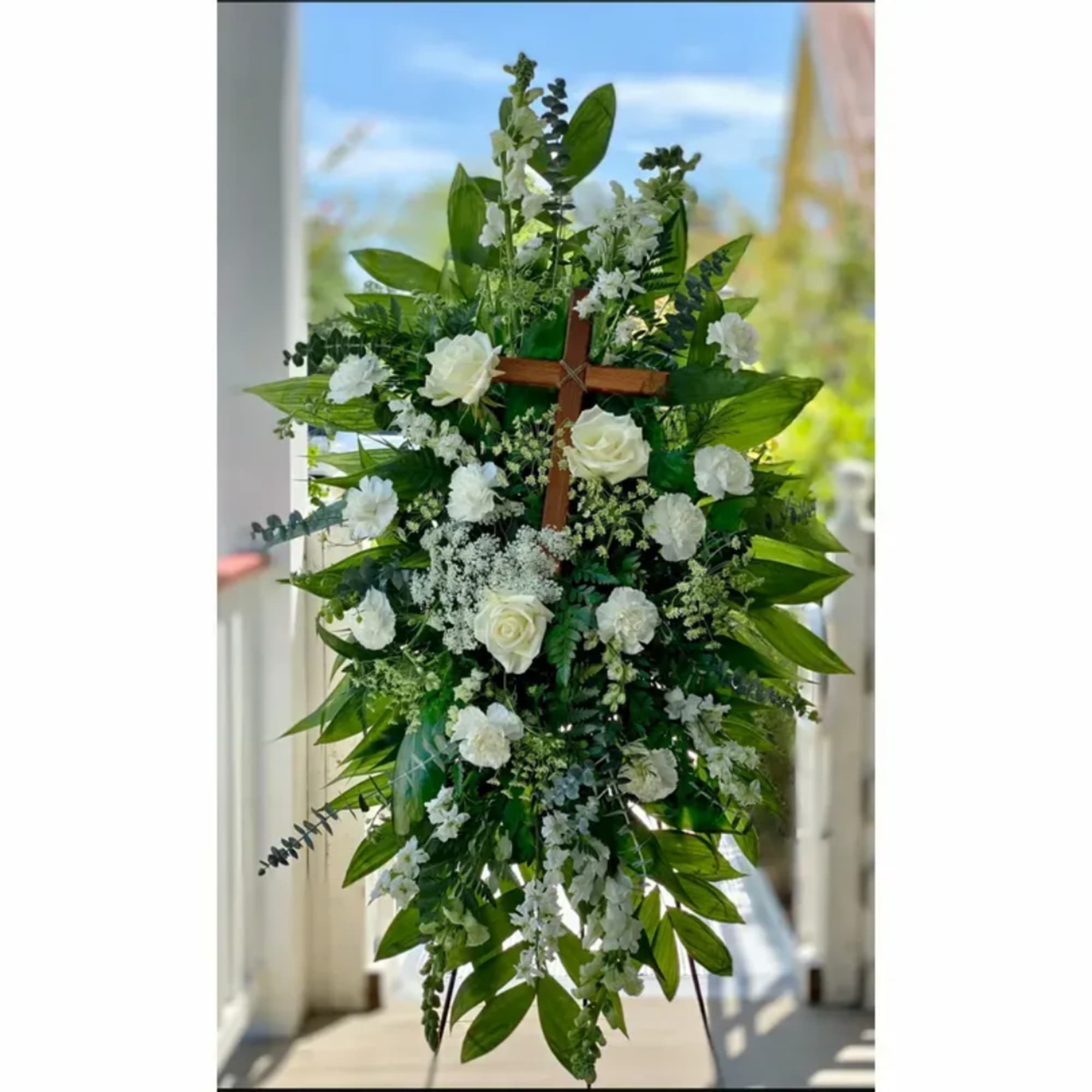 White floral funeral spray on a wooden cross with green foliage