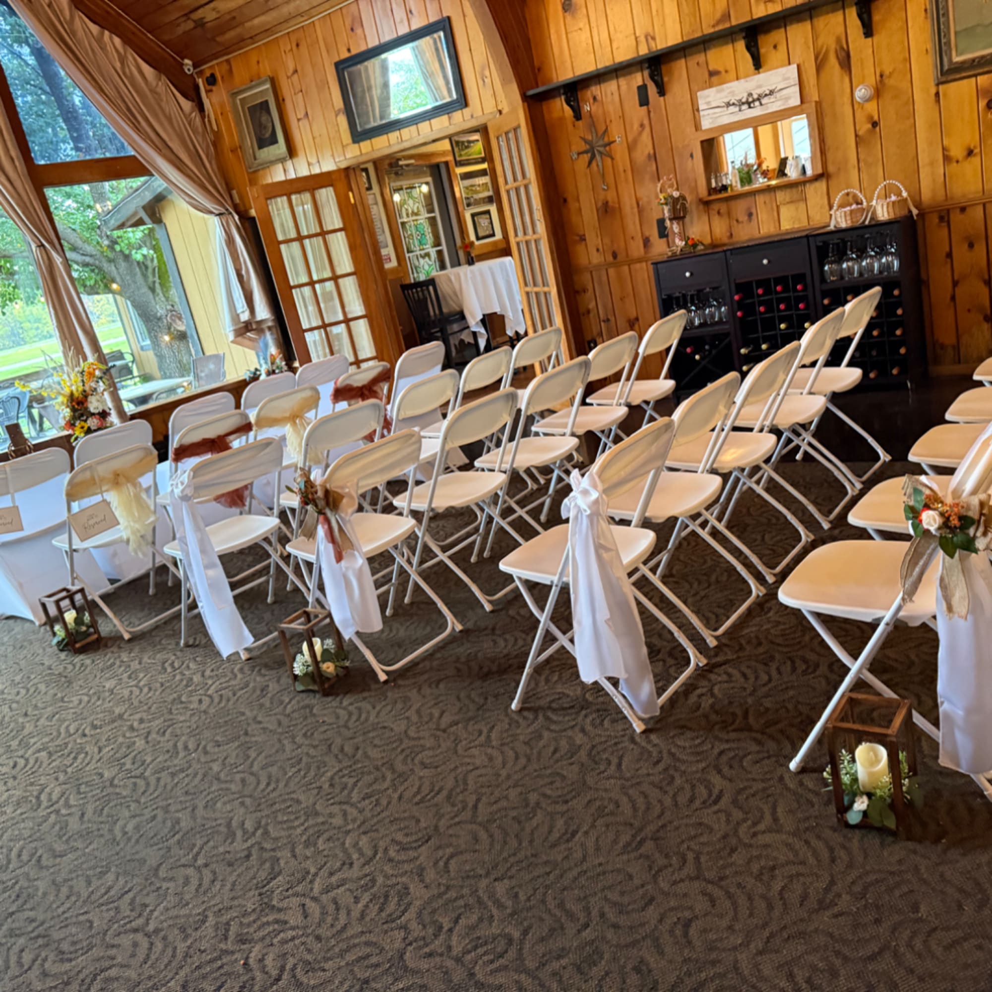 Wedding ceremony setup with white chairs, floral accents, and lanterns in a wood-paneled room.