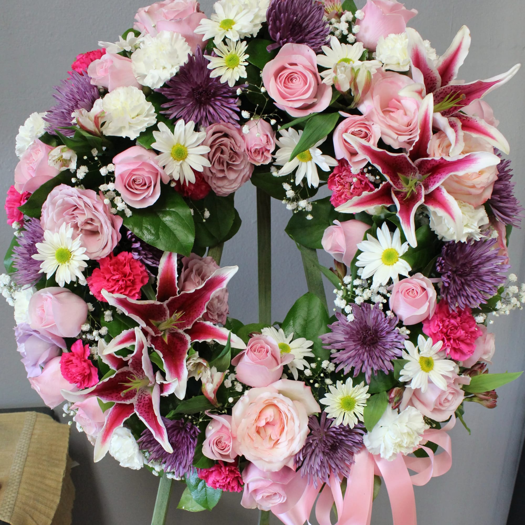 Standing wreath of pink roses, lilies, white daisies, carnations, and purple mums with a pink ribbon on an easel