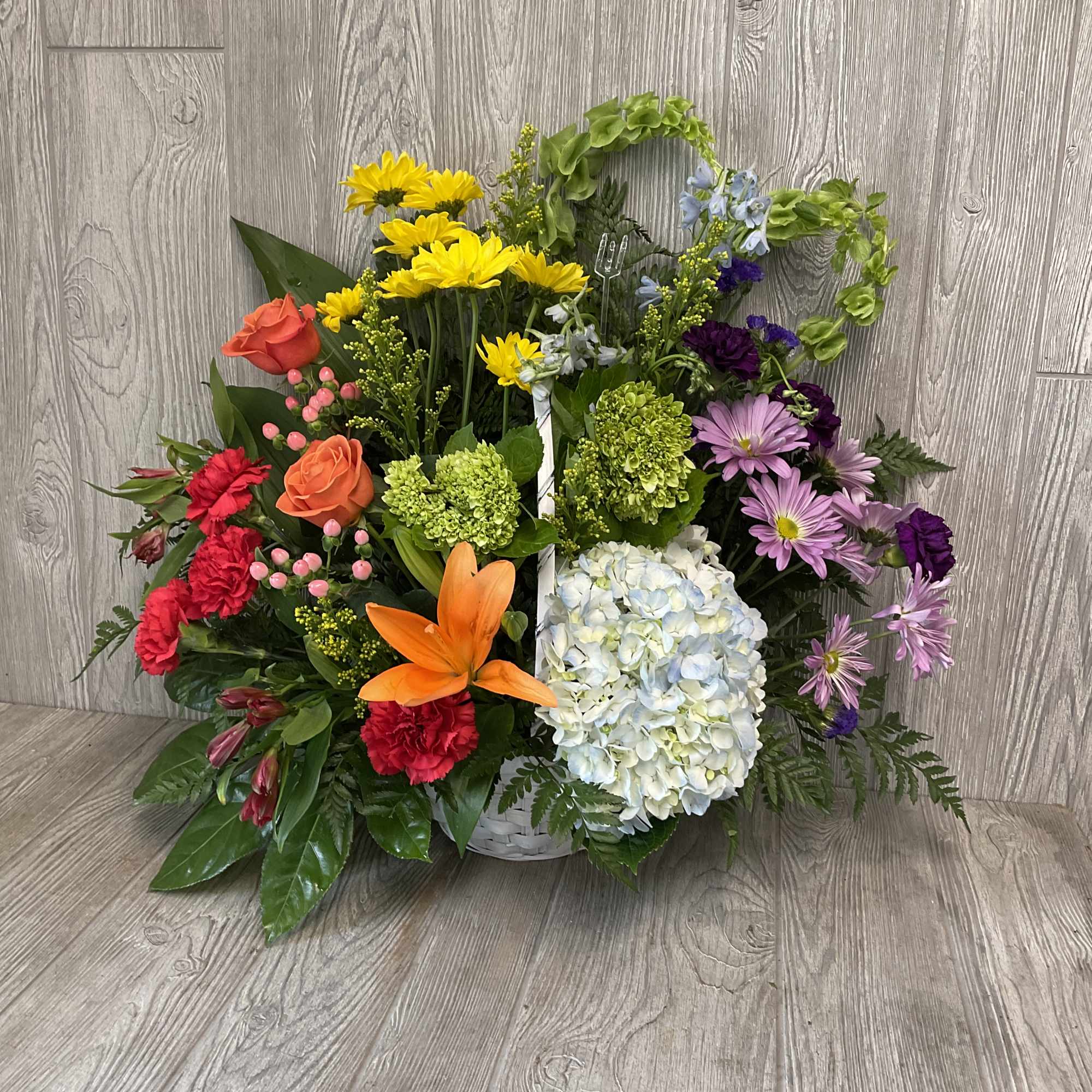 Colorful mixed flower arrangement with lilies, roses, daisies, and hydrangeas in a white basket.