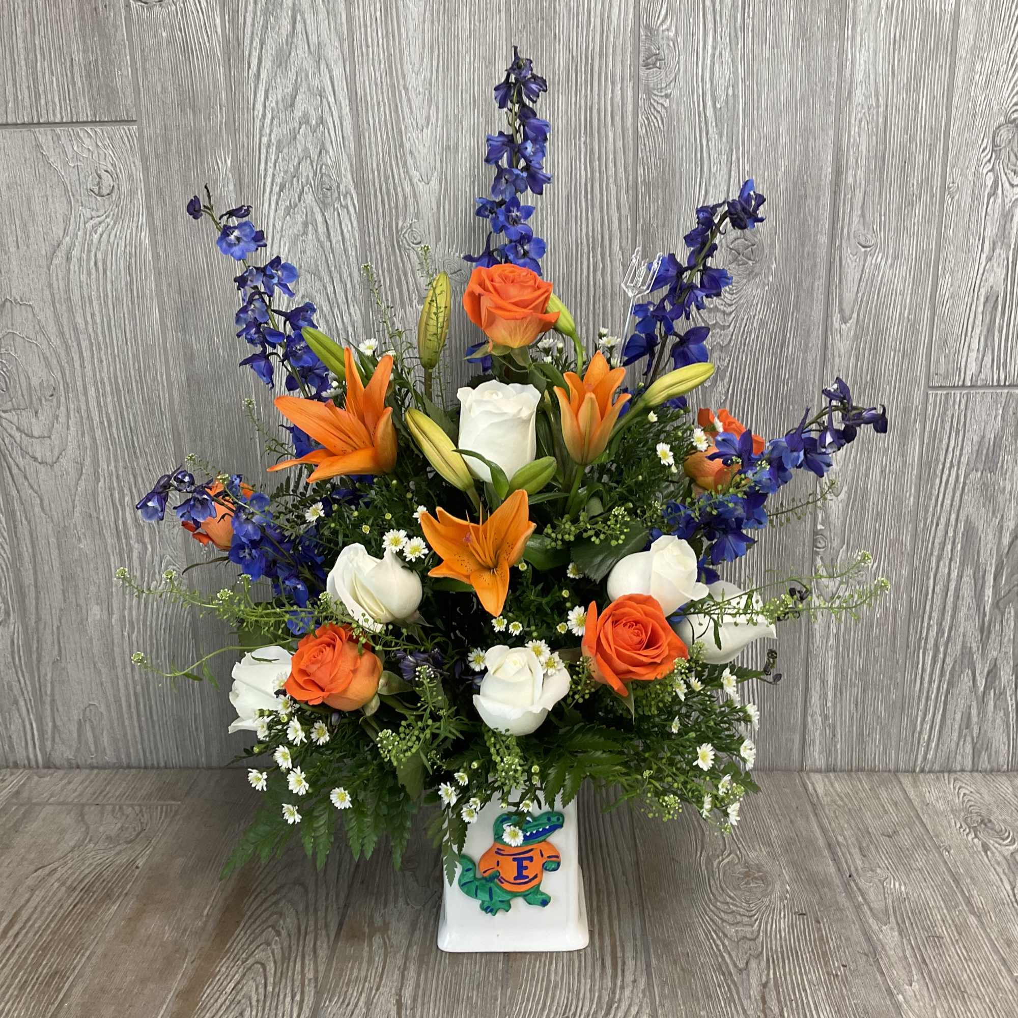 Orange and white roses with lilies in a white vase, accented by blue flowers