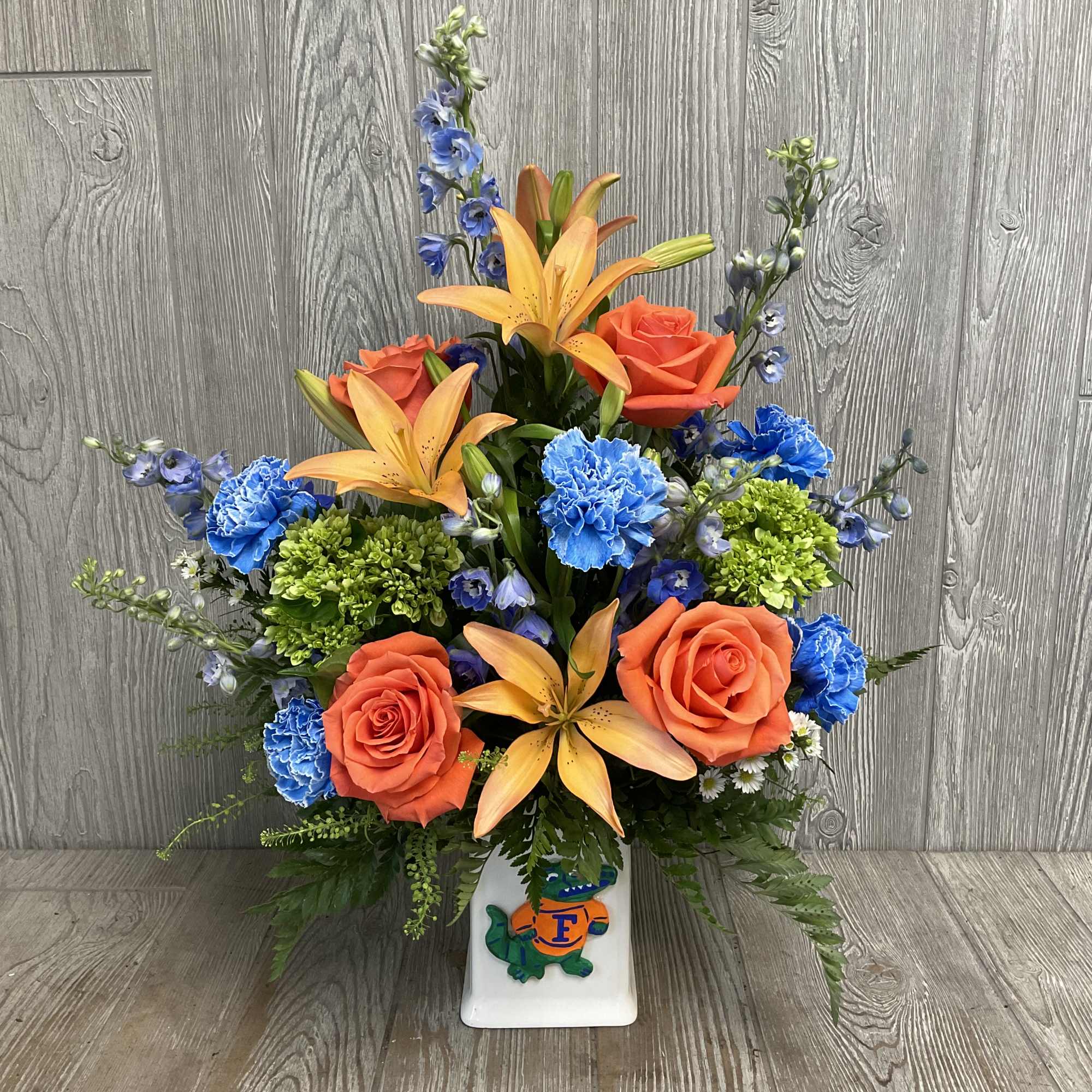 Orange roses and lilies with blue and green accents in a white vase