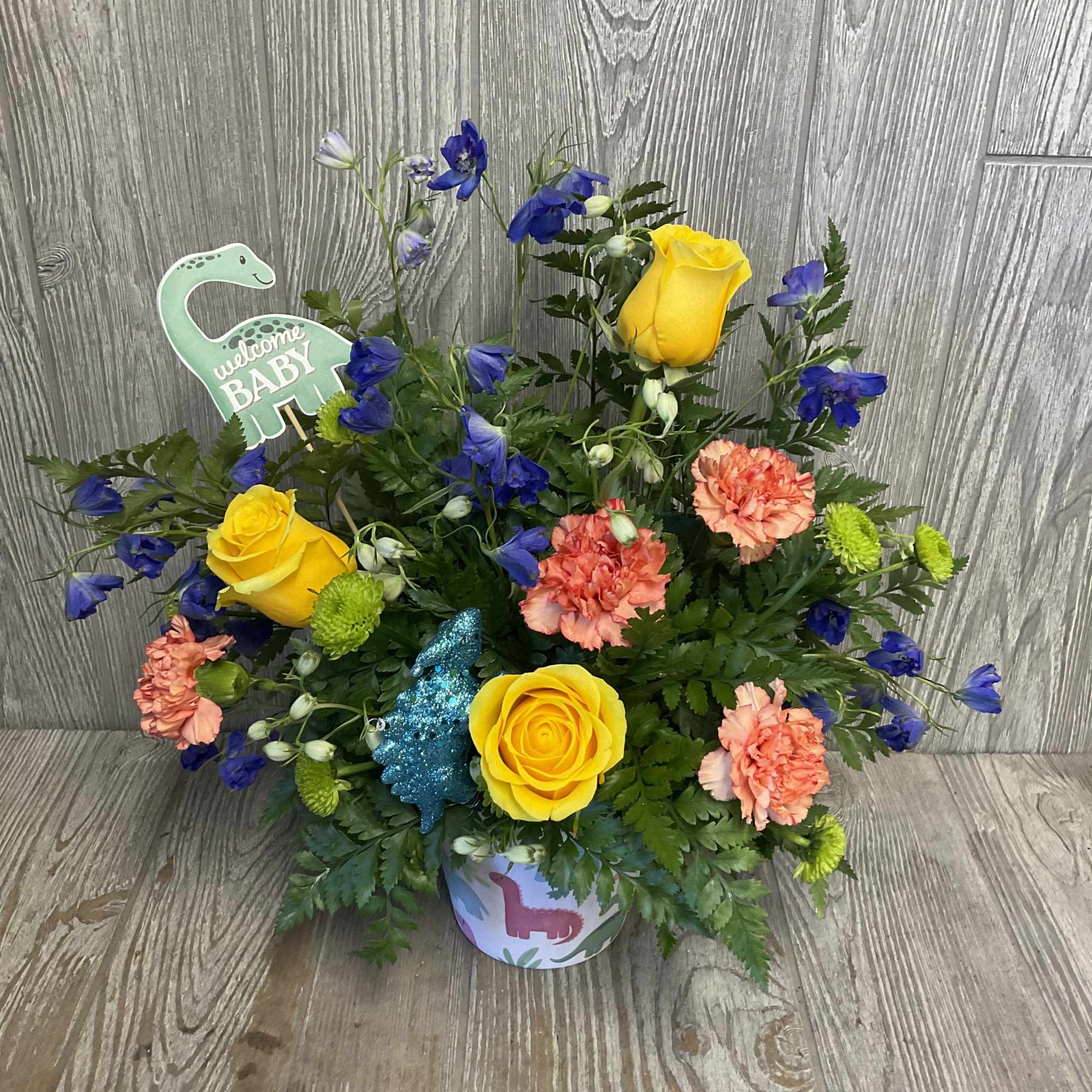 Bouquet of yellow roses, blue flowers, and peach carnations in a baby-themed container