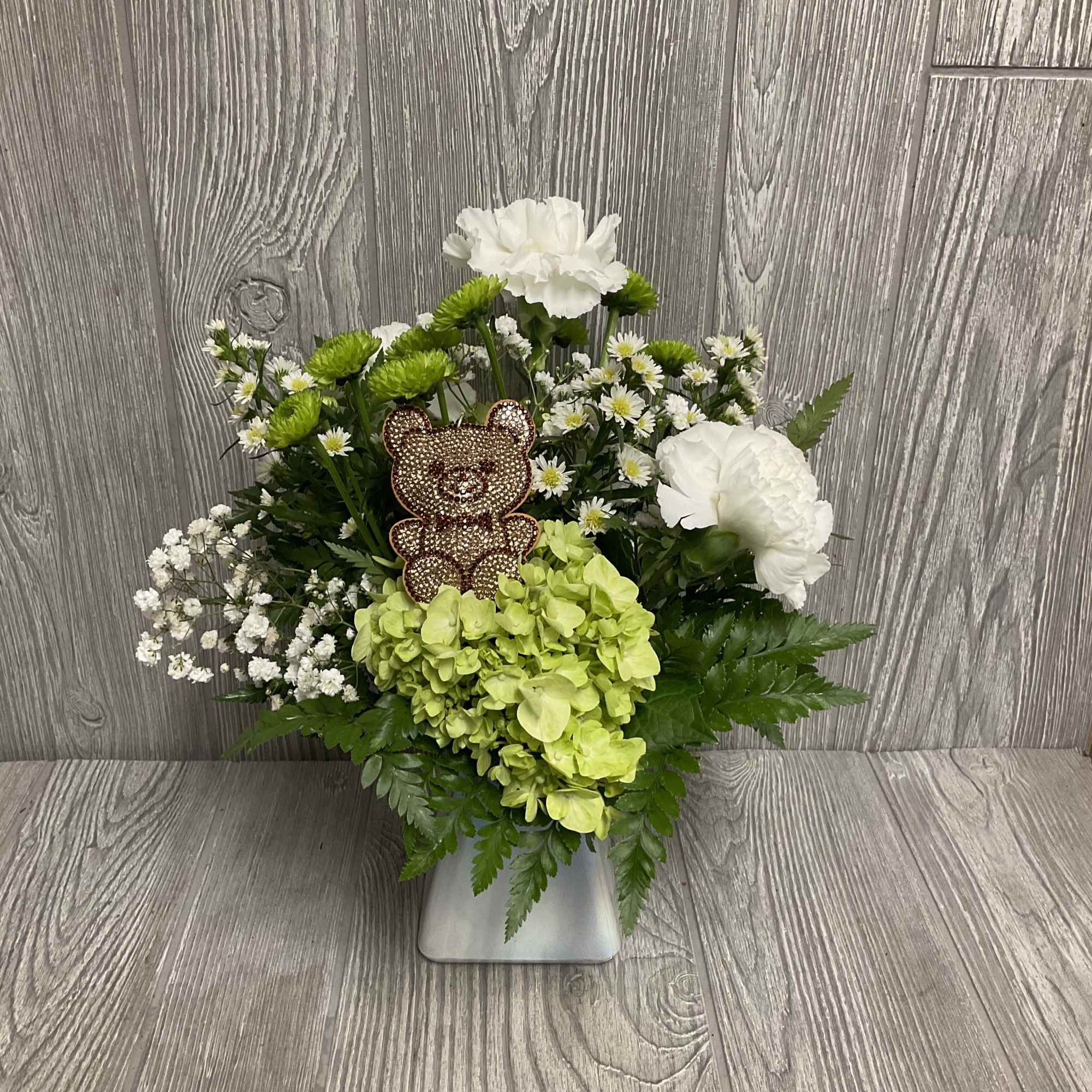 White and green floral arrangement in a silver vase with a teddy bear pick