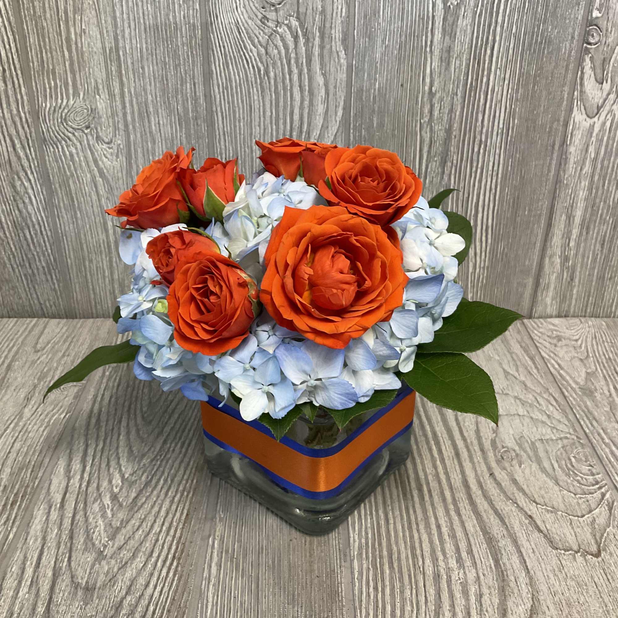 A 4X4 square vase with blue hydrangea and orange roses.  Vase