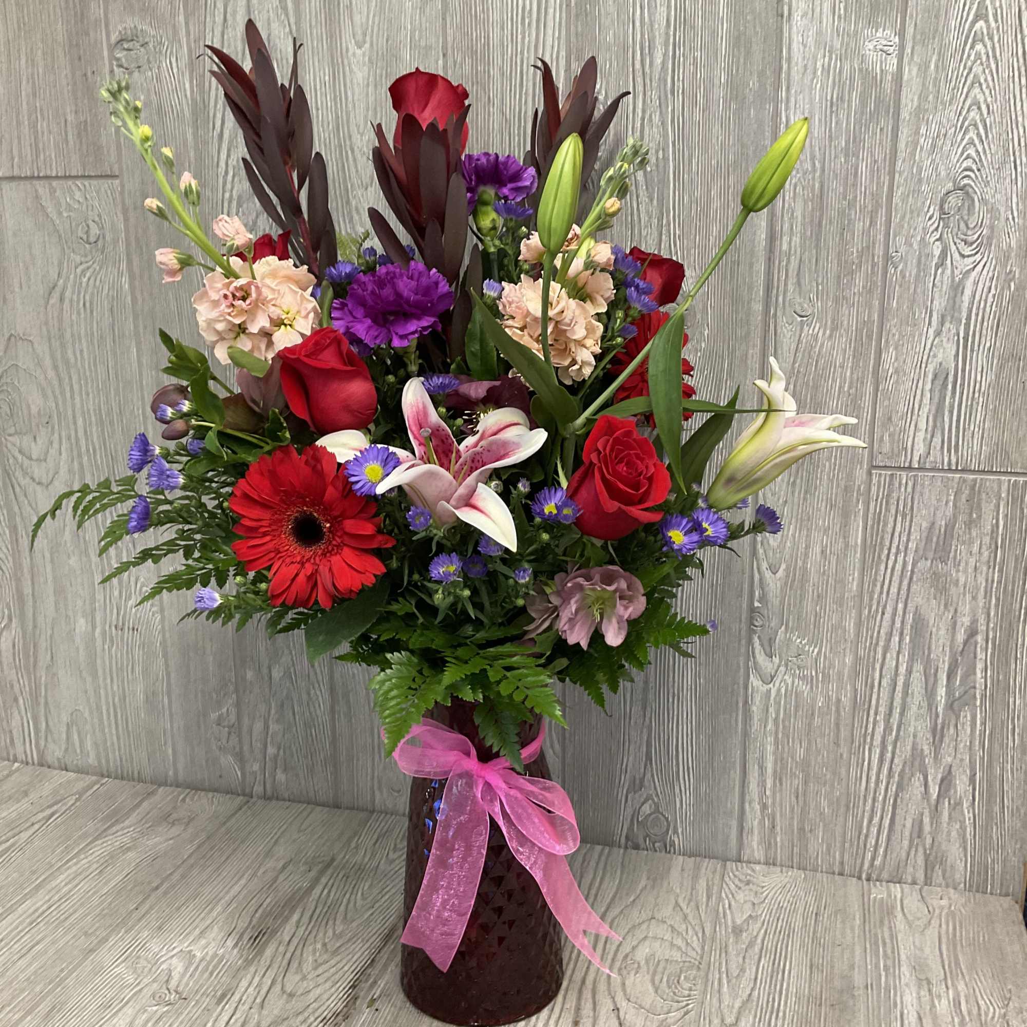 Mixed bouquet of red, pink, purple, and white flowers in a tall vase with a pink ribbon.