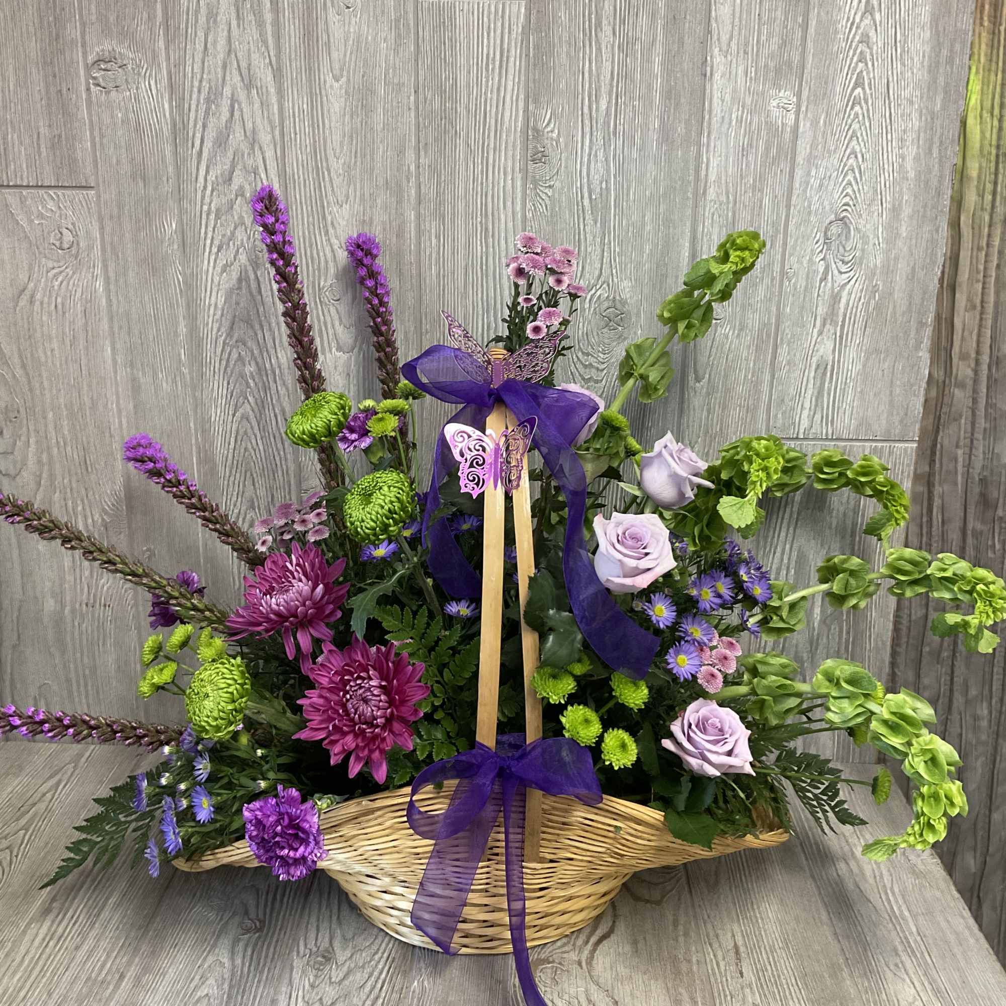 Purple and green floral arrangement in a wicker basket with a ribbon bow