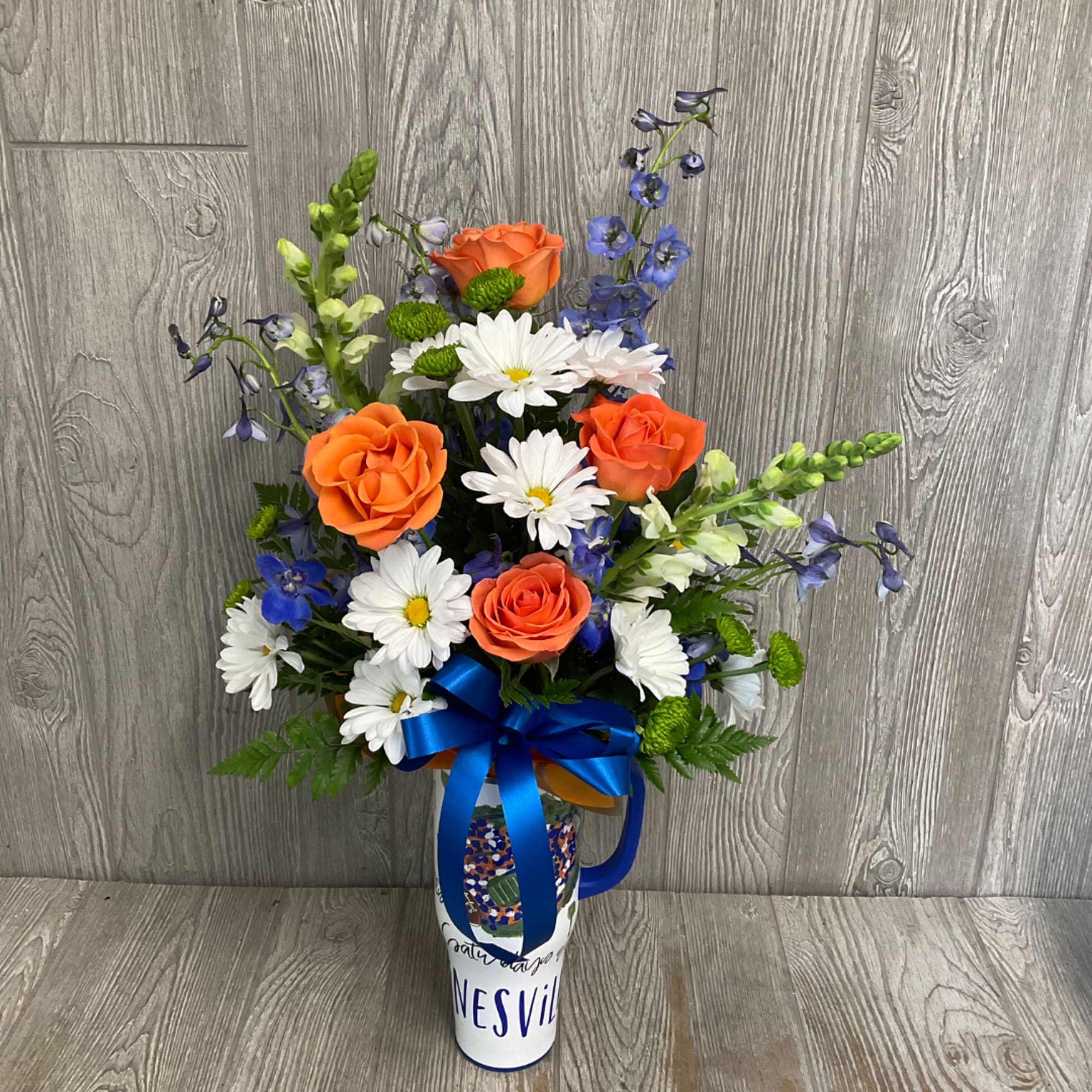 Orange roses and white daisies in a decorated vase with a blue ribbon