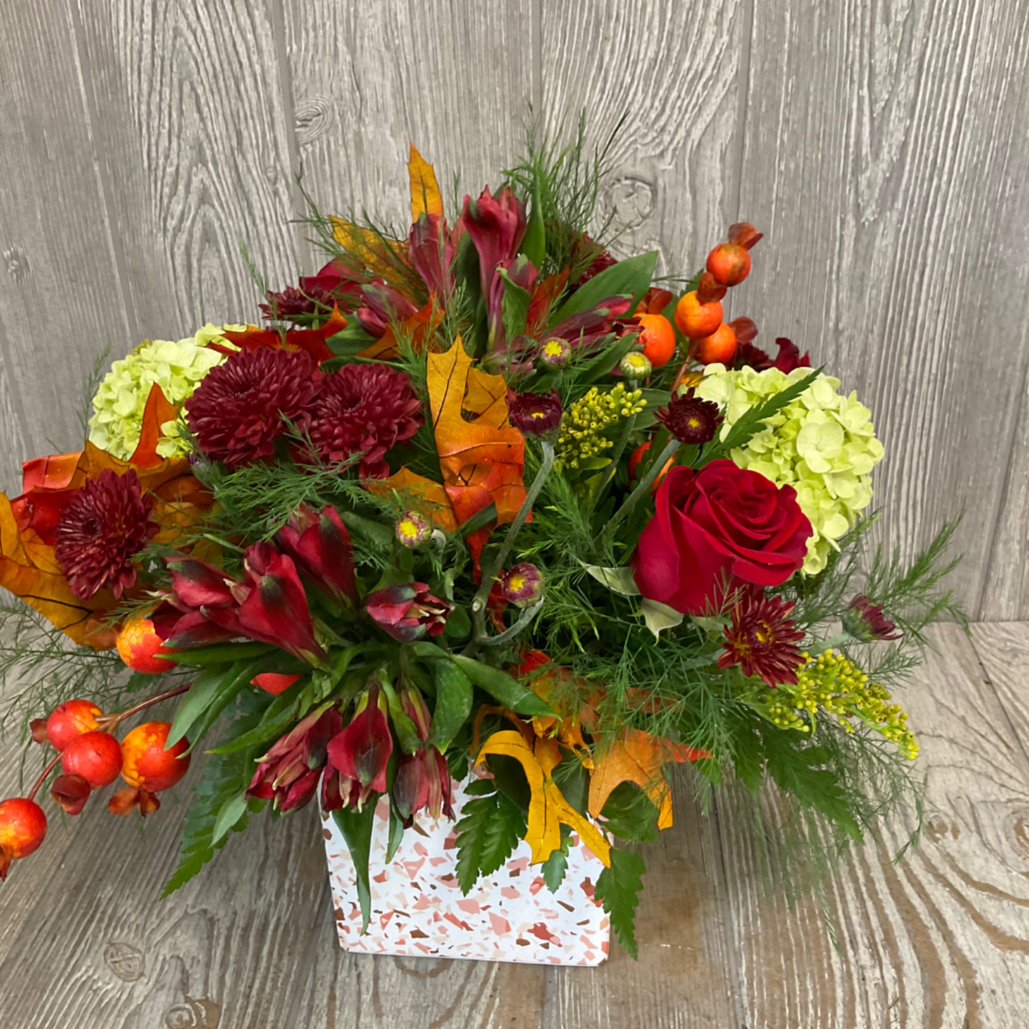 Autumn floral arrangement with red roses, burgundy blooms, and pale green hydrangeas in a patterned box.