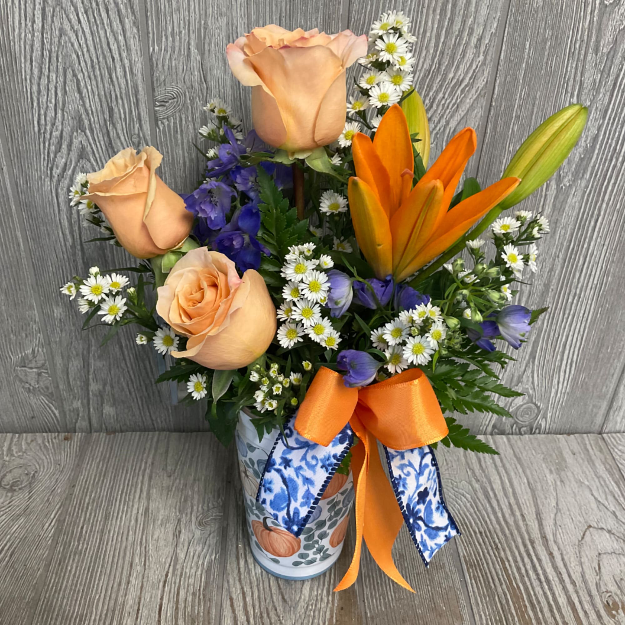 Peach roses, orange lilies, and purple flowers in a patterned vase with ribbon