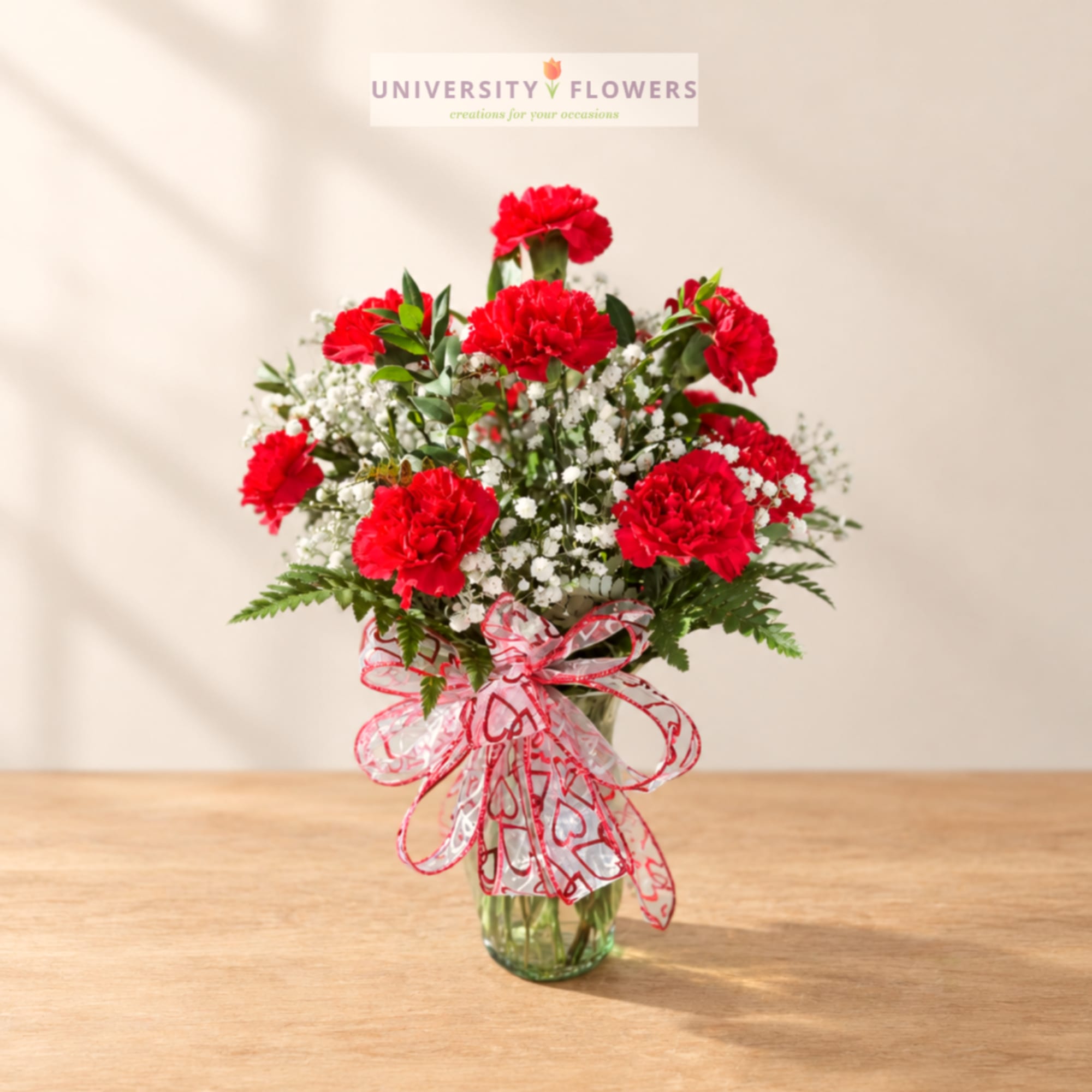 Red carnations and white baby's breath in a glass vase with a heart ribbon