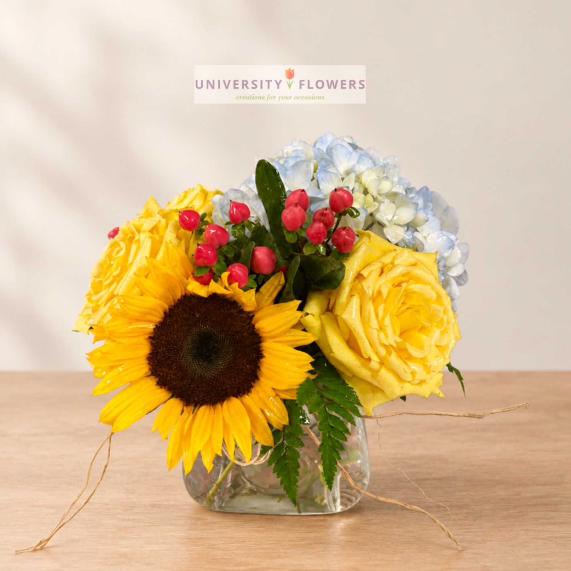 Sunflower and rose arrangement in a clear glass vase with blue hydrangeas