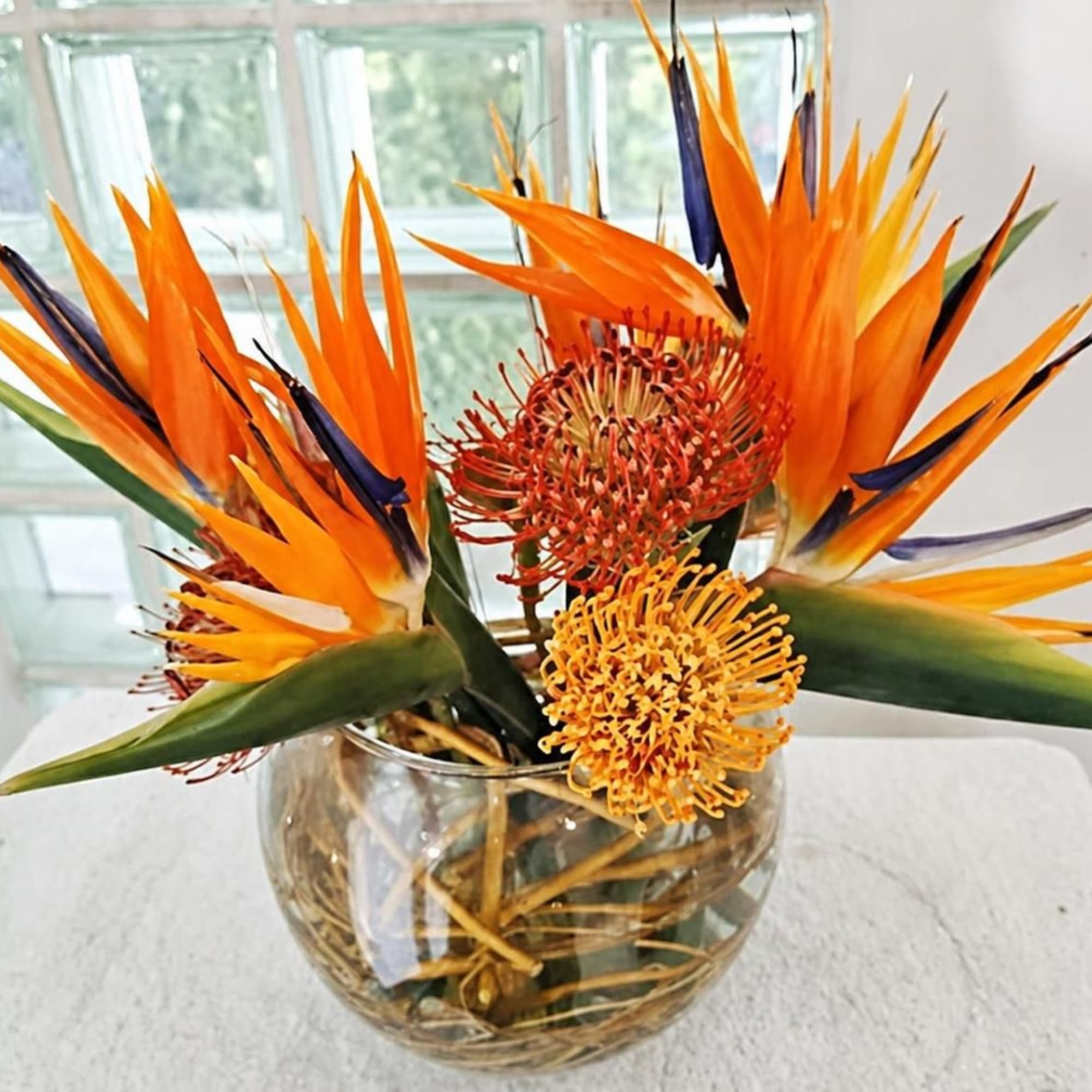 Tropical flower arrangement with orange bird-of-paradise and pincushion blooms in a glass bowl