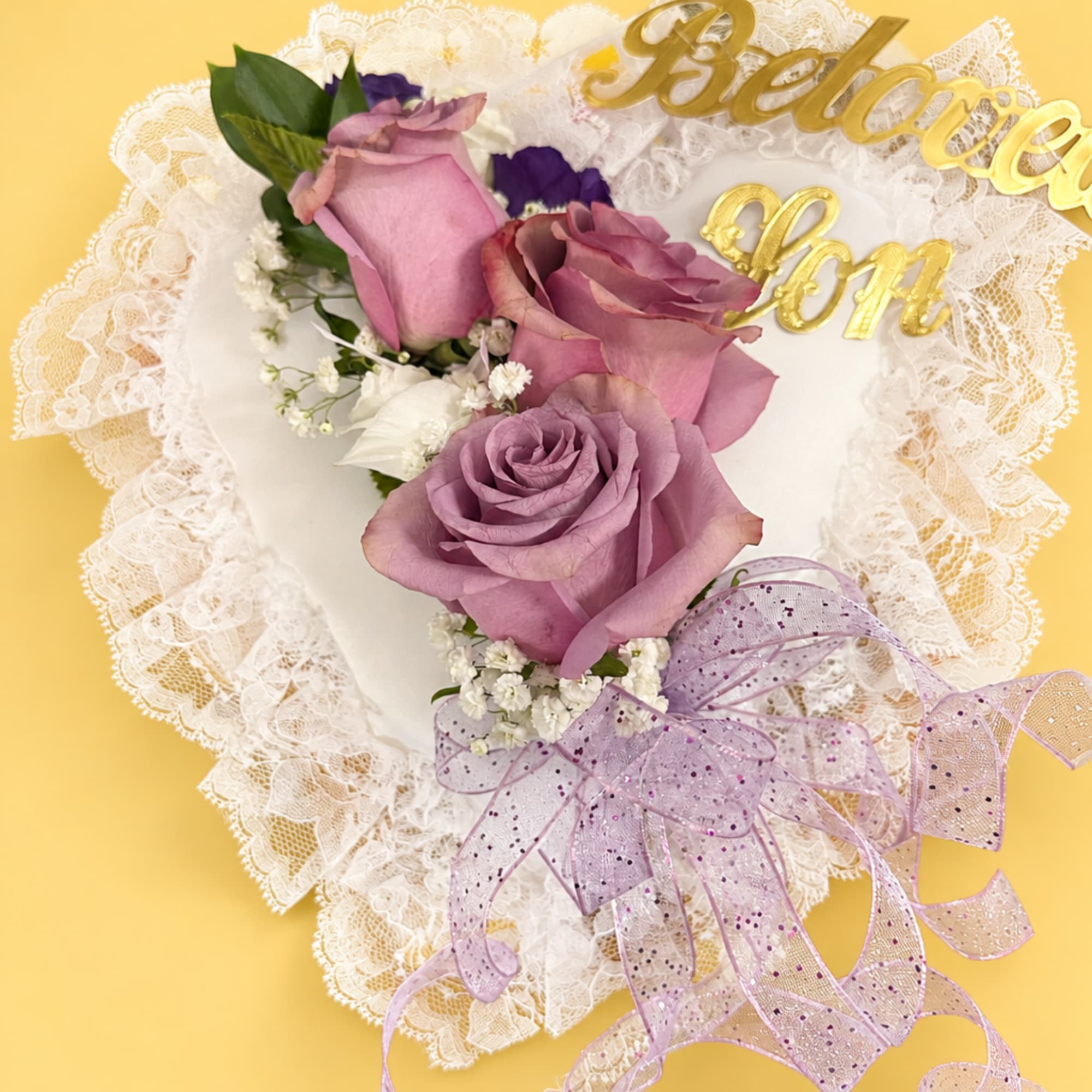 Lavender roses arranged on a lace heart with a gold "Believe" topper