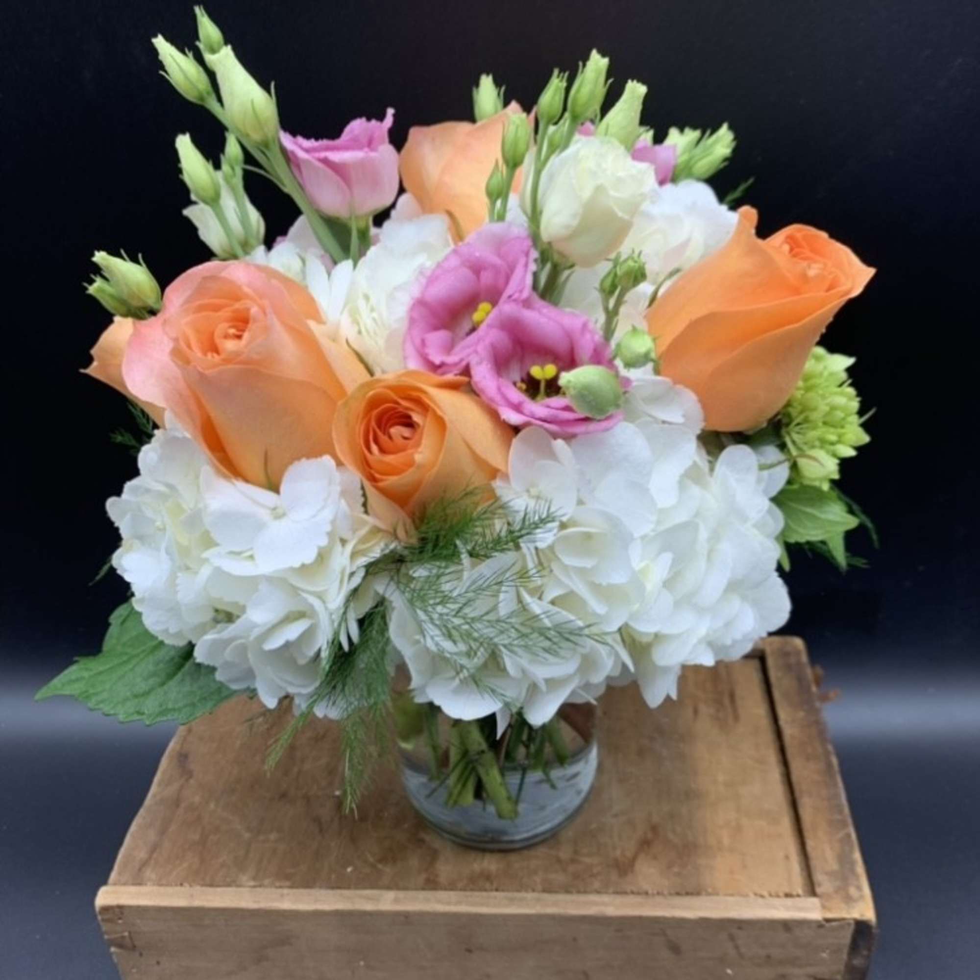 Short arrangement of peach roses, white hydrangeas, and pink lisianthus in a clear glass vase.