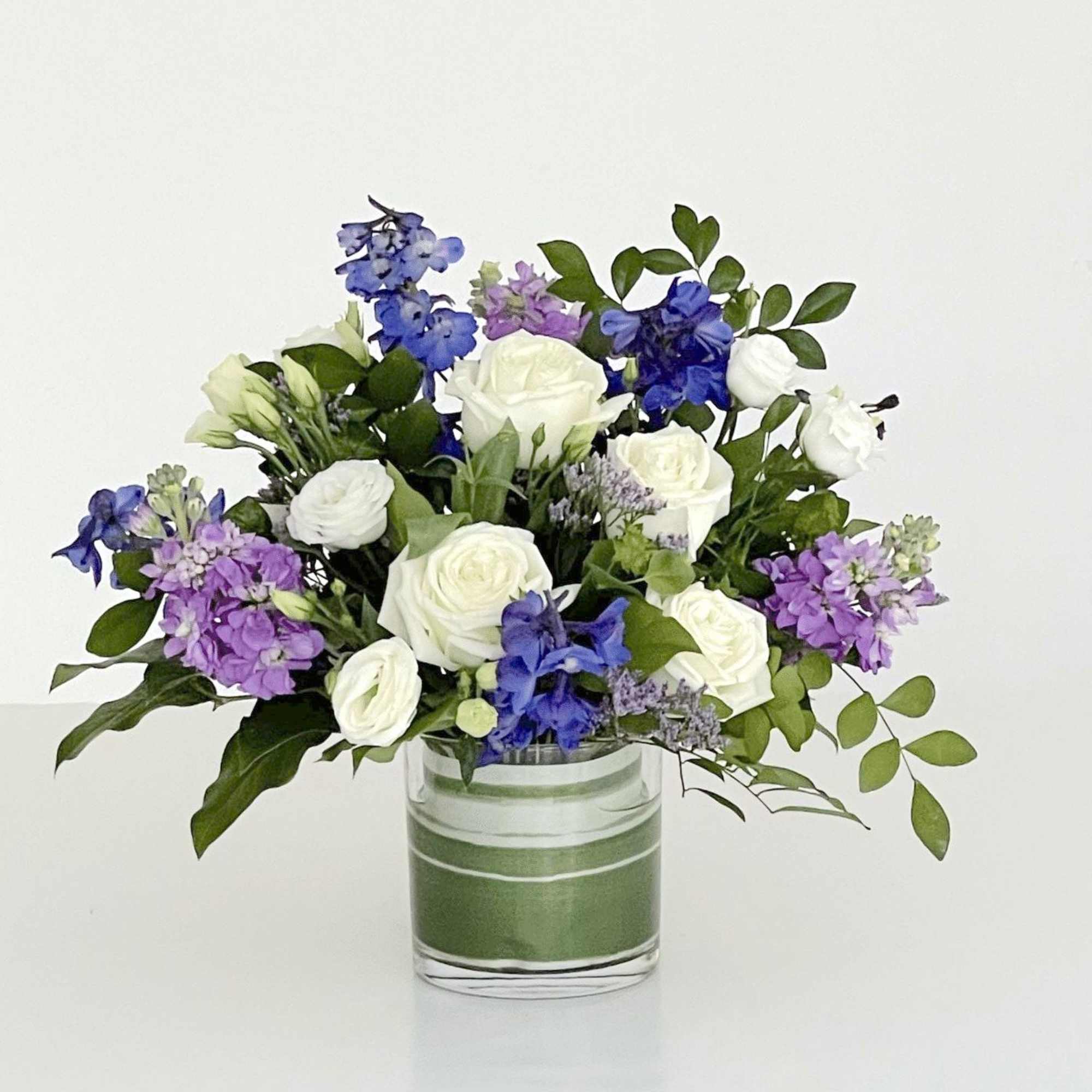 A low leaf-lined cylinder filled with roses, Stock, Lisianthus and Delphinium -