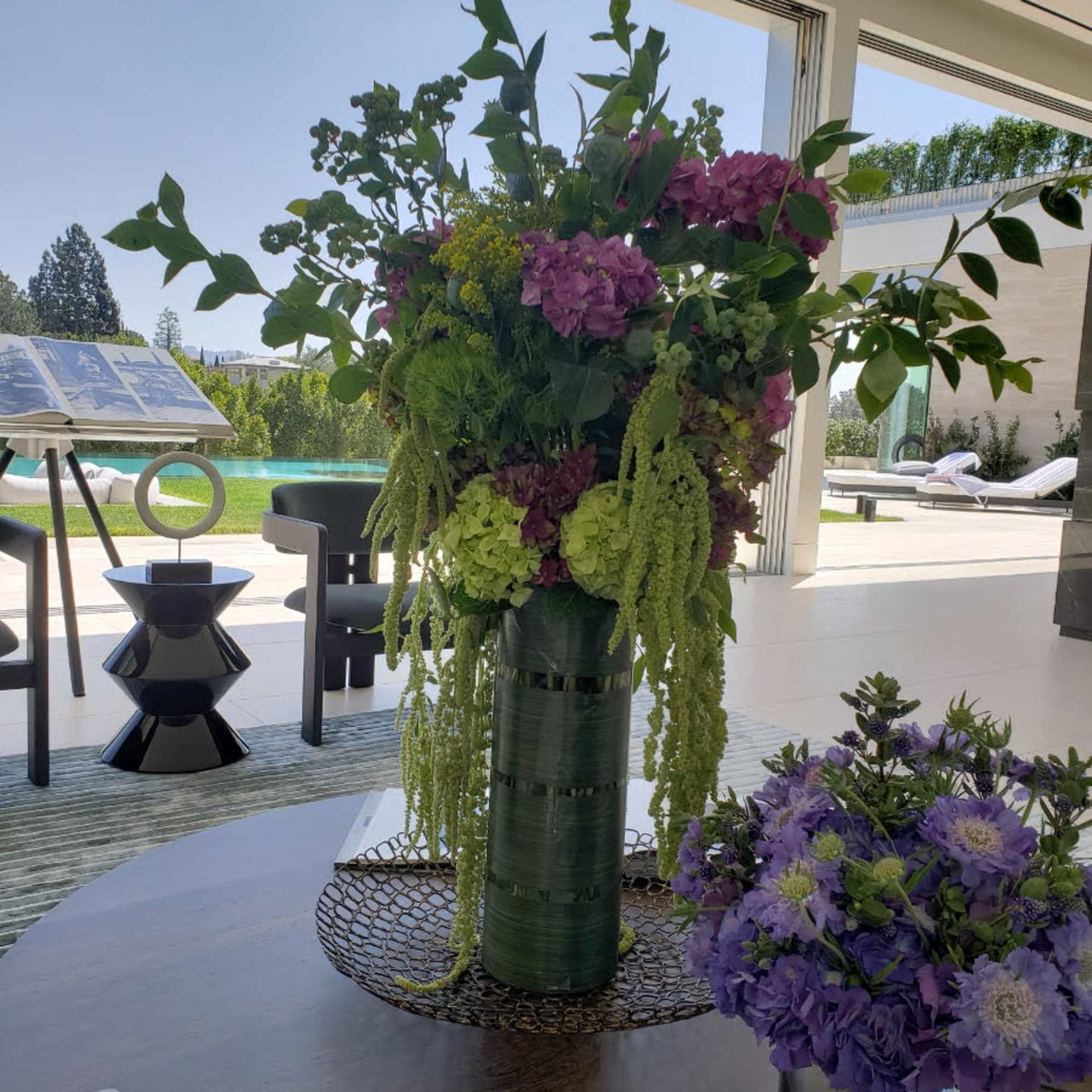 Tall floral arrangement with purple blooms in a green vase beside a smaller purple bouquet