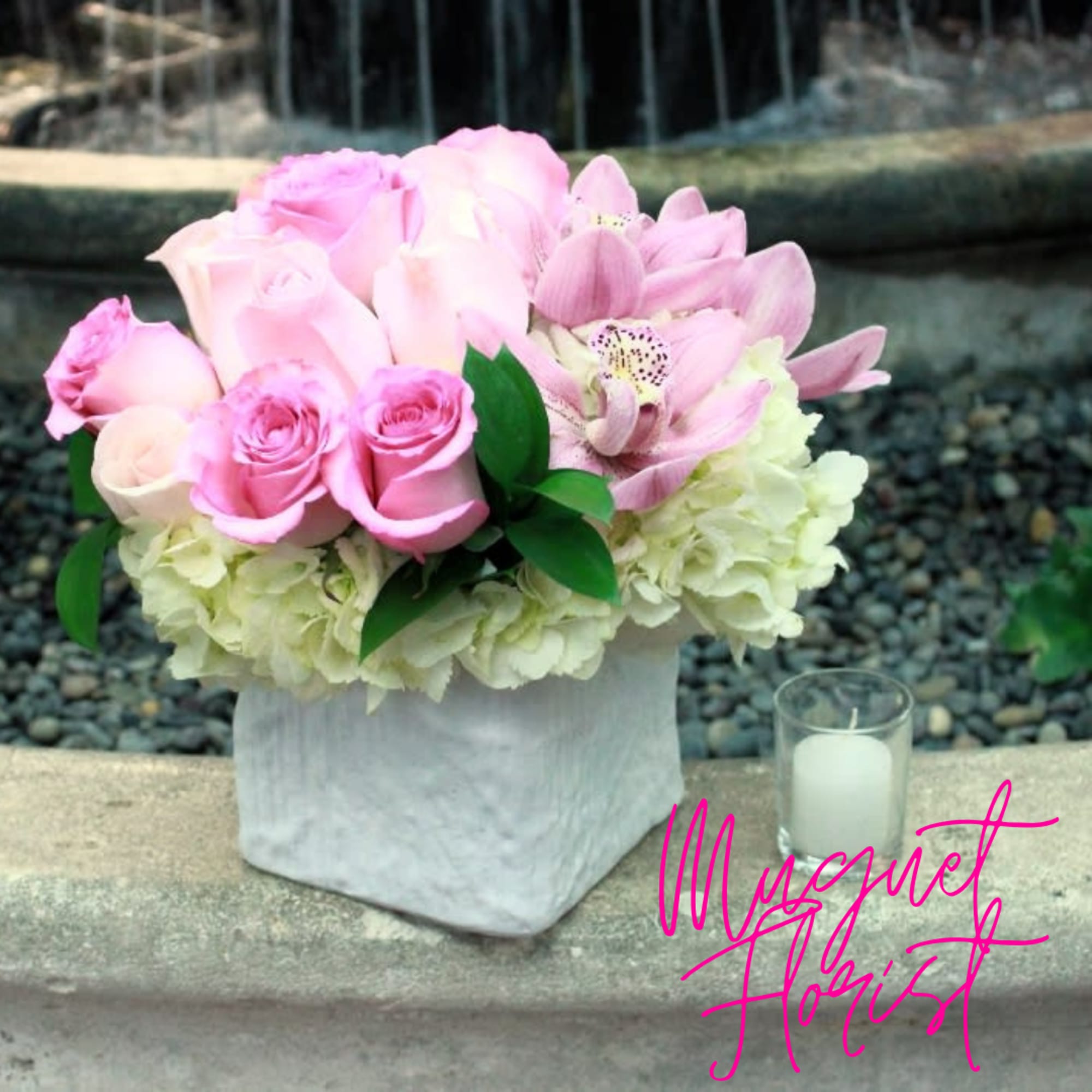 Pink roses and orchids arranged in a white square vase beside a small candle.