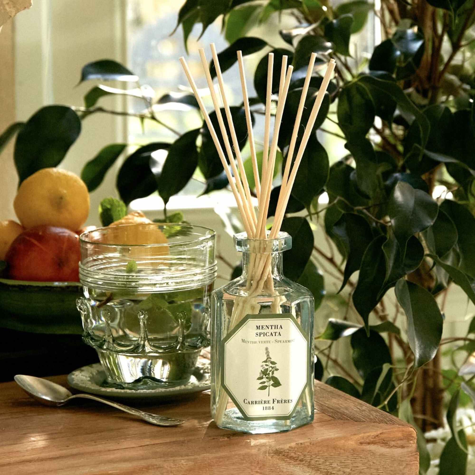 Glass reed diffuser bottle on a table beside a glass cup and fruit bowl