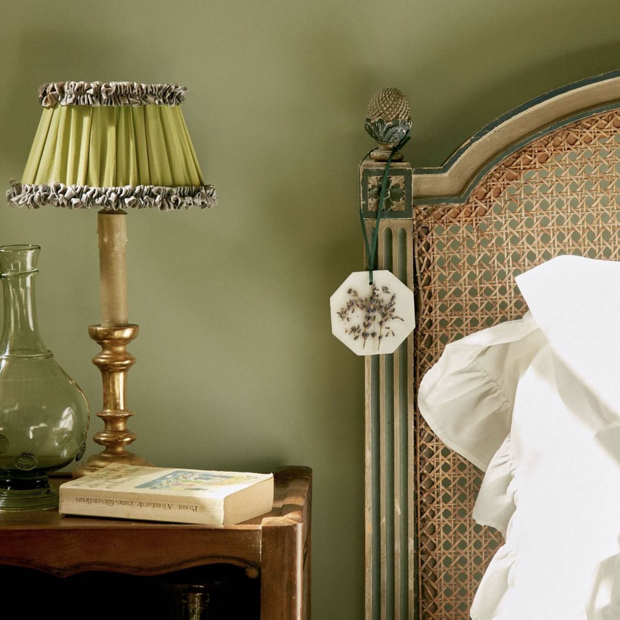 A bedside table with a lamp, glass bottle, book, and woven headboard