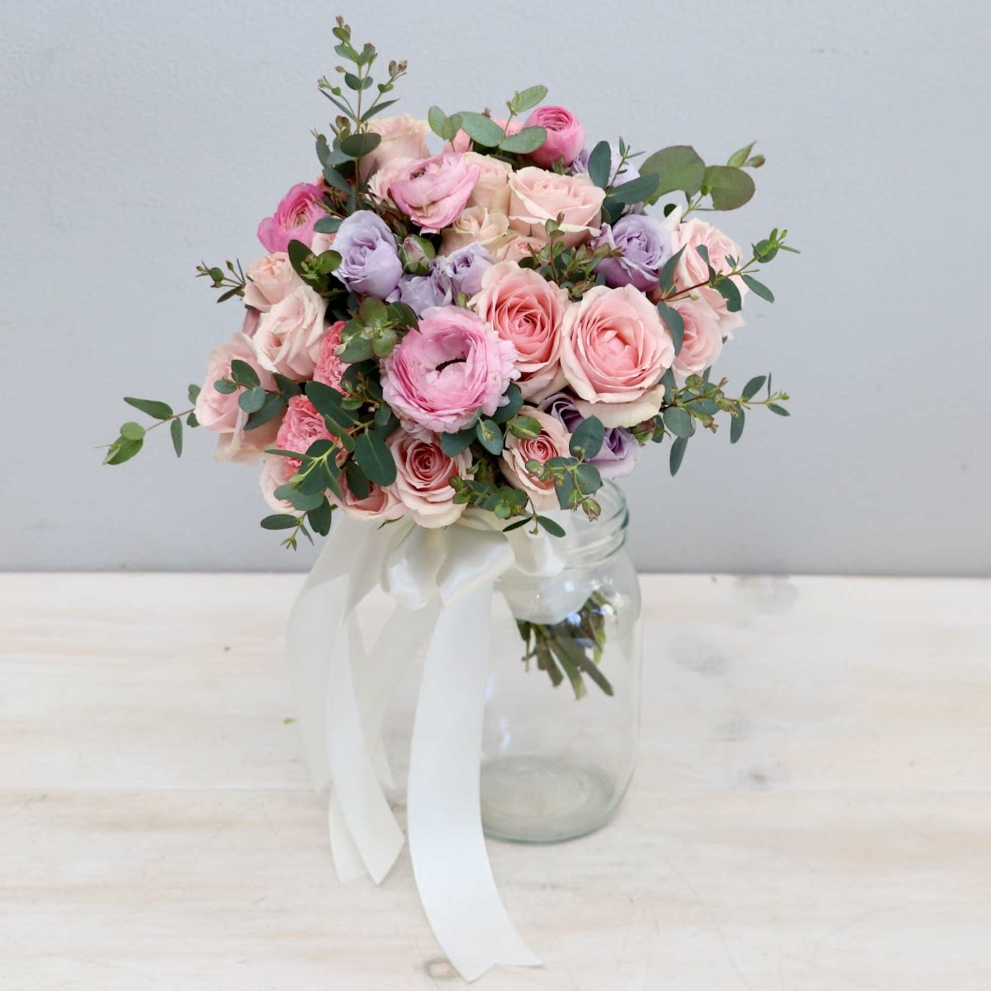 Round bouquet of pink and lavender roses and ranunculus in a clear jar with white ribbon