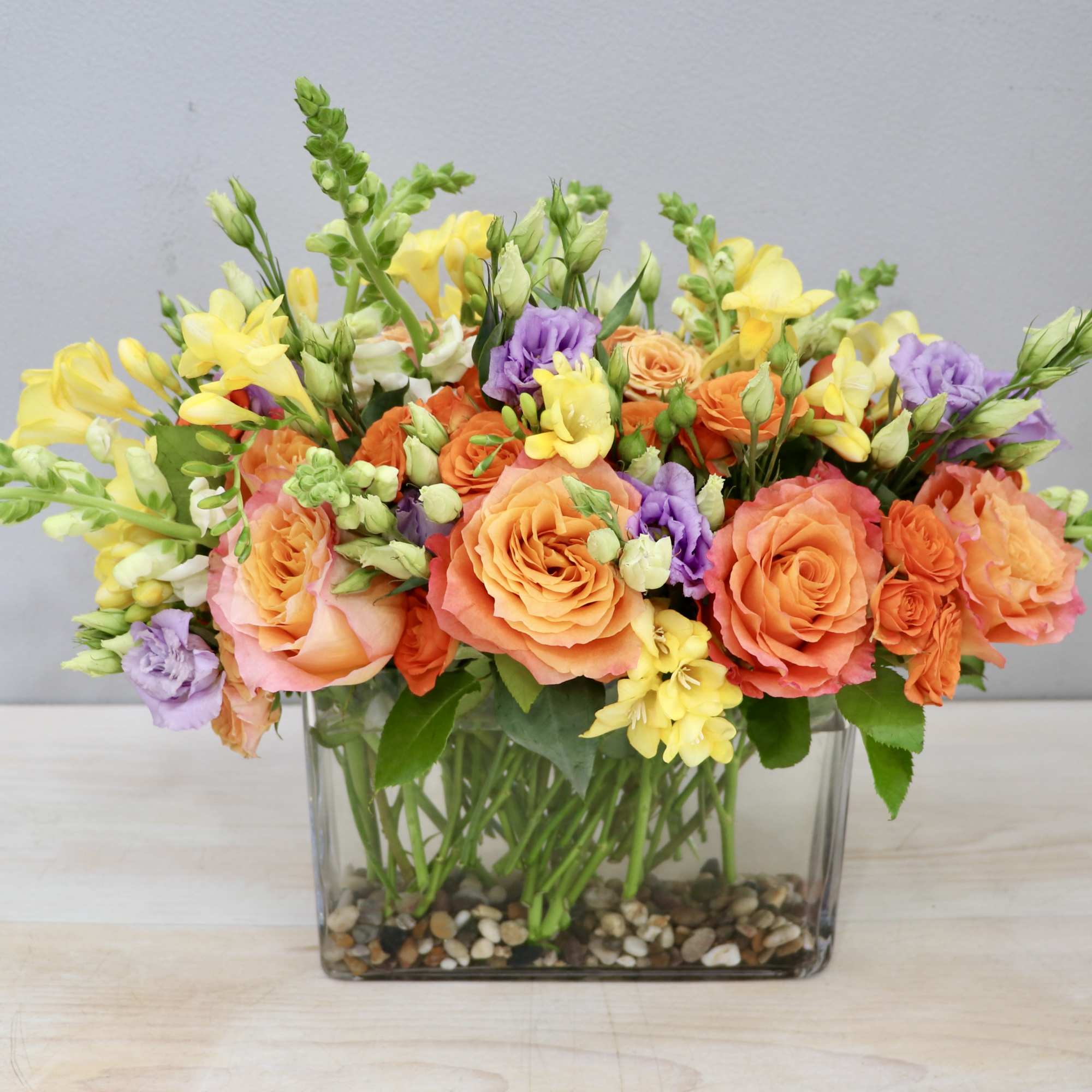 Low rectangular arrangement of orange roses, yellow flowers, and purple blooms in a glass vase with pebbles