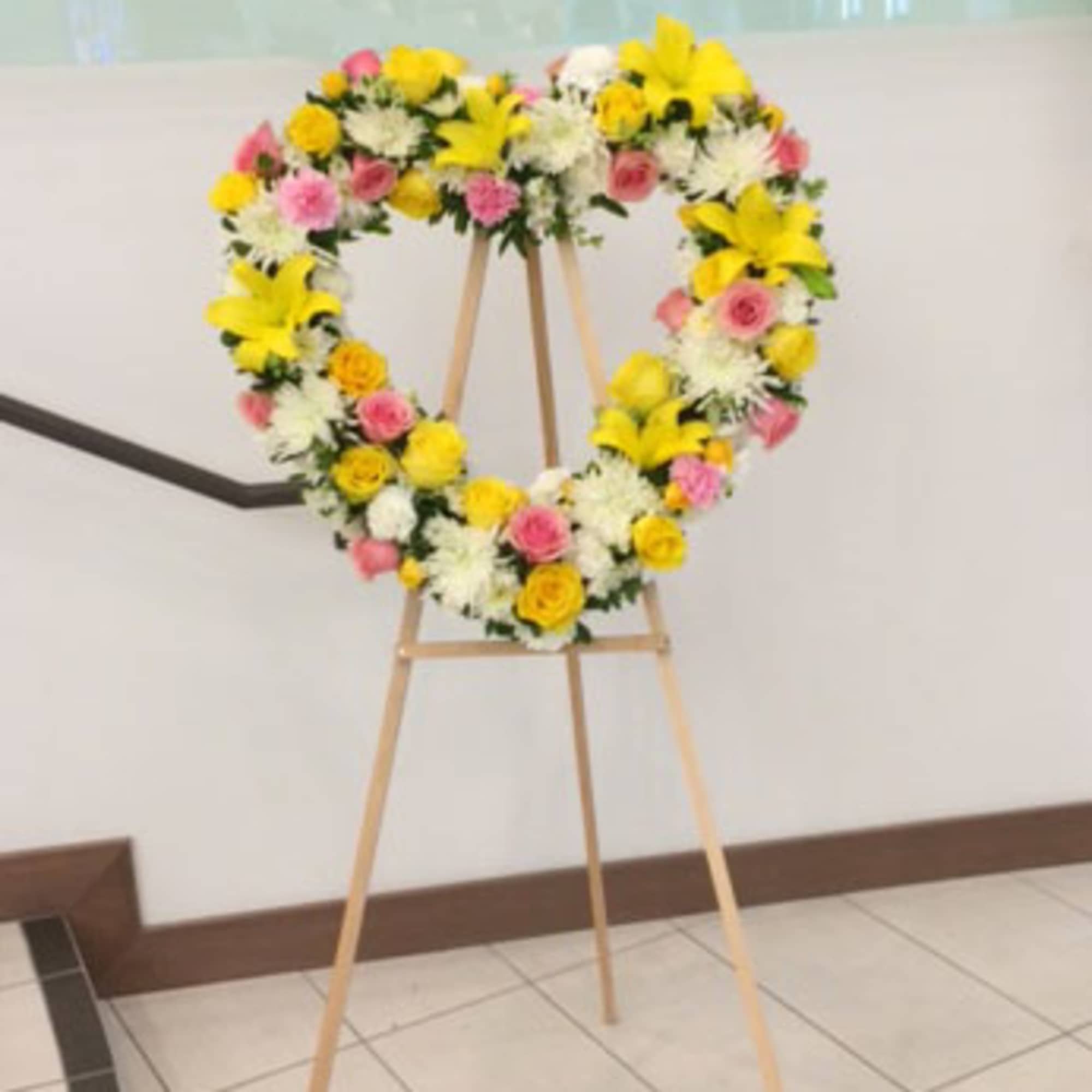 Heart-shaped floral wreath on a wooden easel with yellow, pink, and white flowers