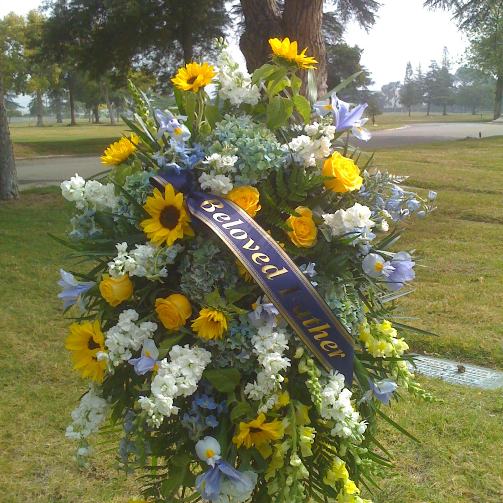 Large funeral spray with sunflowers, yellow roses, and blue flowers on an easel