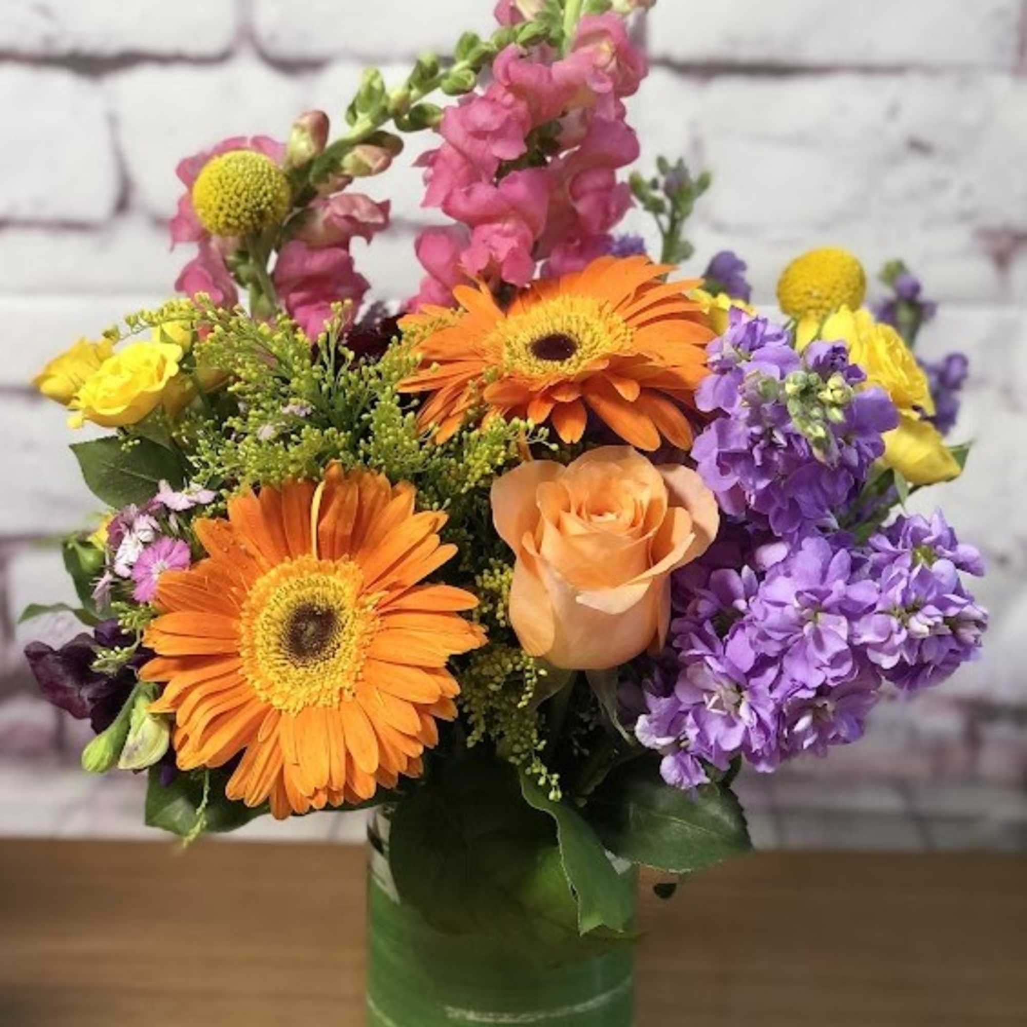 Mixed bouquet with orange gerbera daisies, roses, and purple blooms in a glass vase