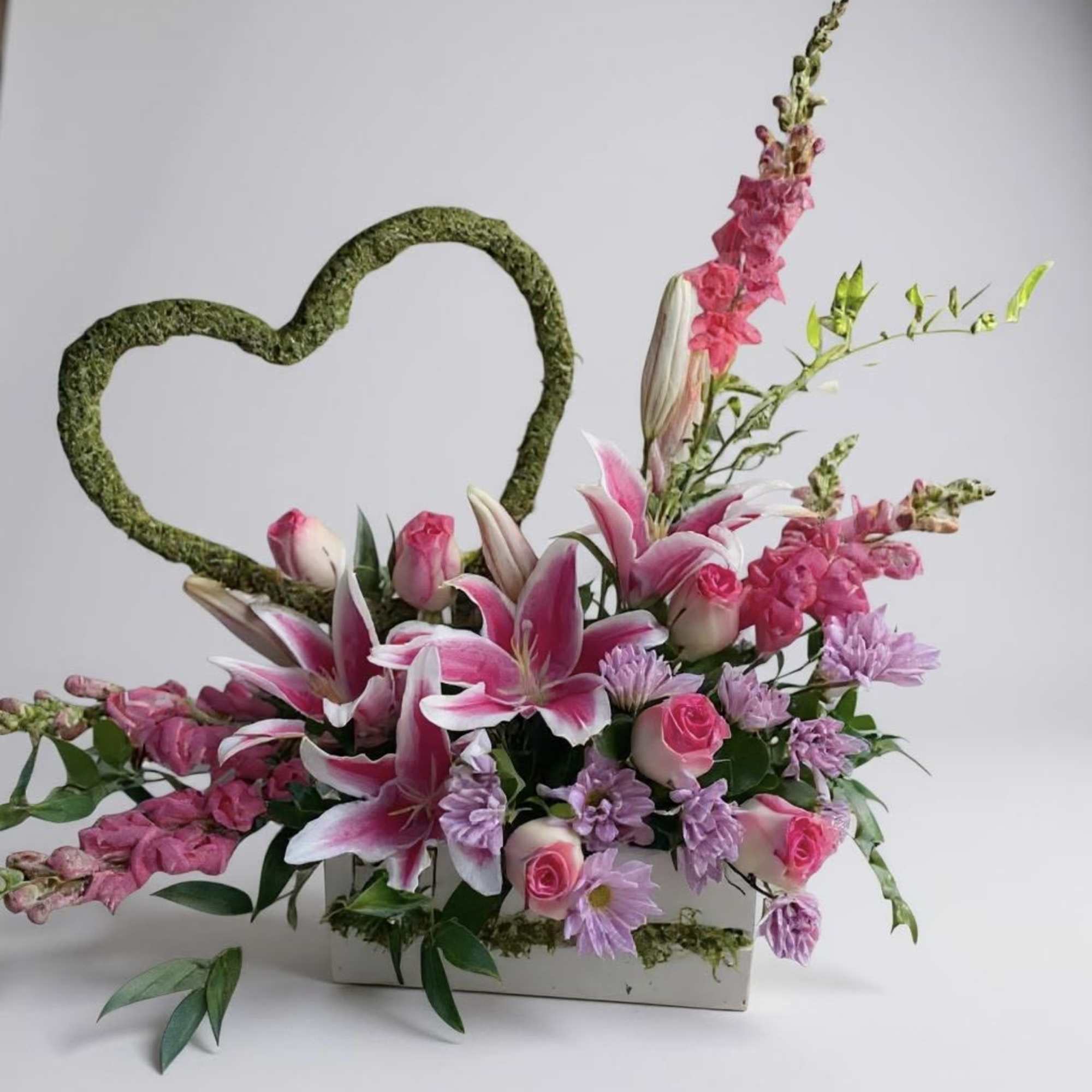 Pink and lavender floral arrangement with a moss heart frame in a white container
